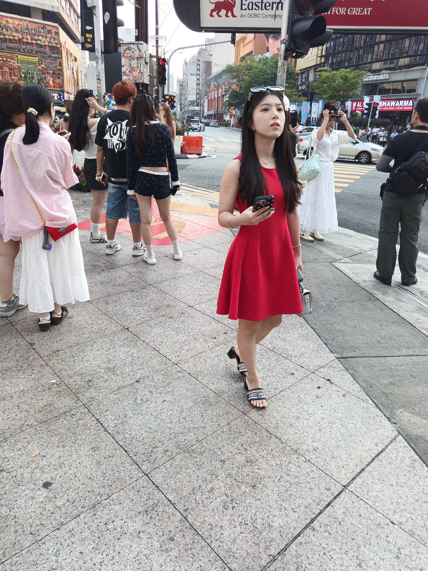 In Kuala Lumpur, Malaysia, on a city sidewalk, a young woman with long dark hair, wearing a red dress and black slides with "CHRISTIAN" inscribed on them, holds a smartphone. She is walking past a group of people, including a woman in a white dress and a man in a black t-shirt and green pants. In the background, a busy street is visible, with traffic, buildings, and signs for businesses such as "McDonald's" and "Shawarma". The scene is captured from a low angle, emphasizing the woman in the red dress.