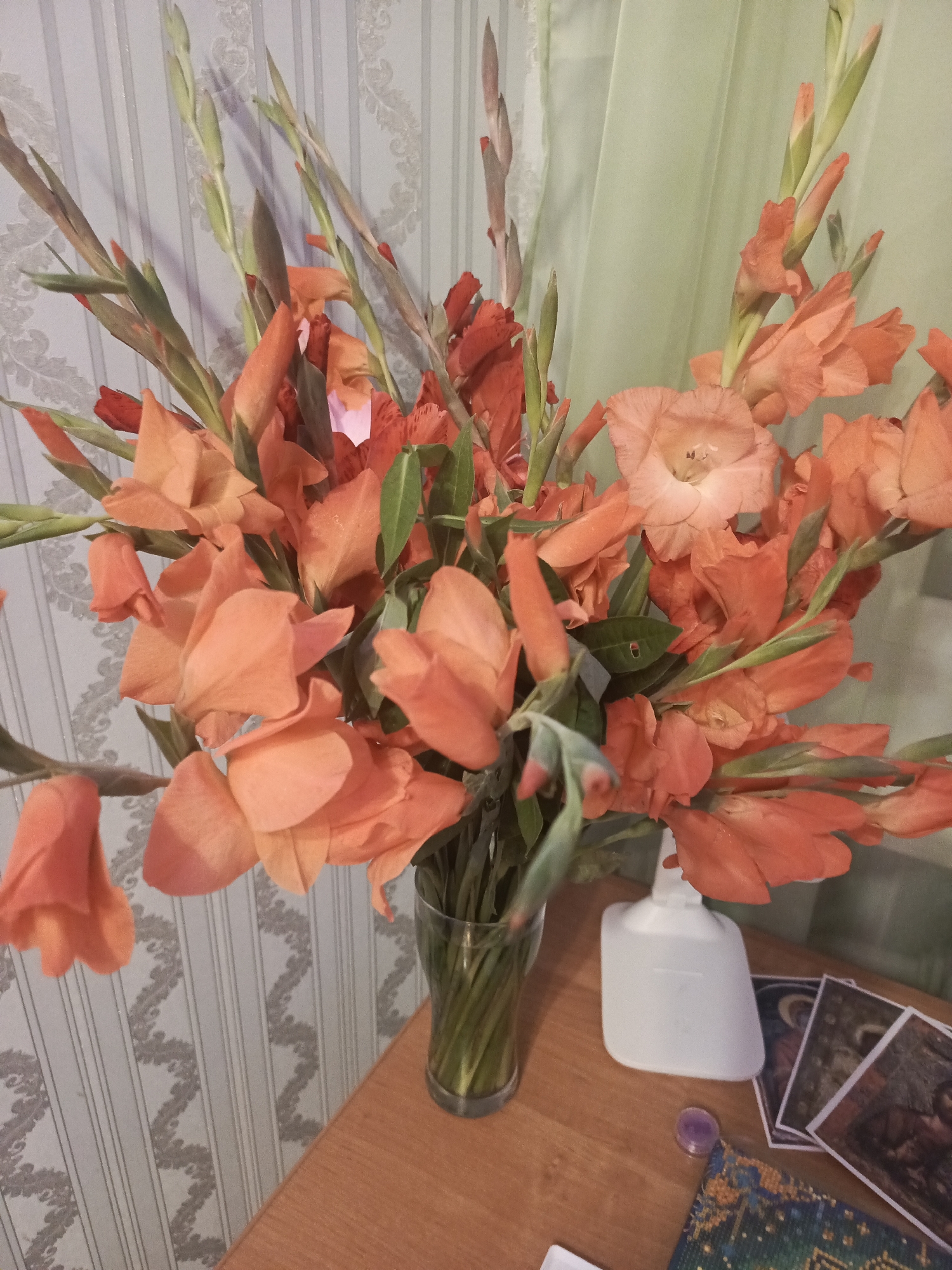 A bouquet of vibrant orange and red gladiolus flowers fills a clear glass vase on a wooden surface. The flowers are arranged in a loose, natural style, with some buds still closed and others in full bloom. The background features a light-colored wallpaper with a subtle striped and damask pattern, and a pale green curtain. To the right of the vase, a white bottle is positioned next to a collection of tarot cards and a small purple object. The scene appears to be an indoor setting, likely a home or a shop, possibly in Nalchik, Russia, given the context provided.