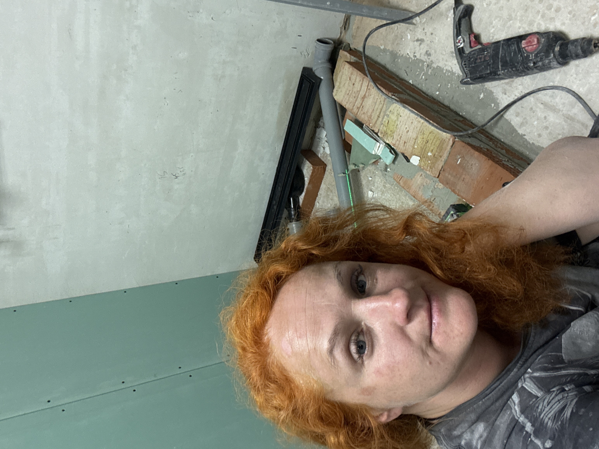 A selfie taken indoors shows a woman with red hair, fair skin, and blue eyes. She is wearing a grey t-shirt with a black and white pattern. In the background, the scene shows construction or renovation work. There is a light green wall, possibly drywall, with screw holes. Above that, there's a section of concrete or plastered wall. Near the top, there's a grey PVC pipe connected to a black rectangular object. Bricks, tools, and construction debris are scattered around, including a black power drill. Based on the prompt, the location is in Chaikovsky, Russia.