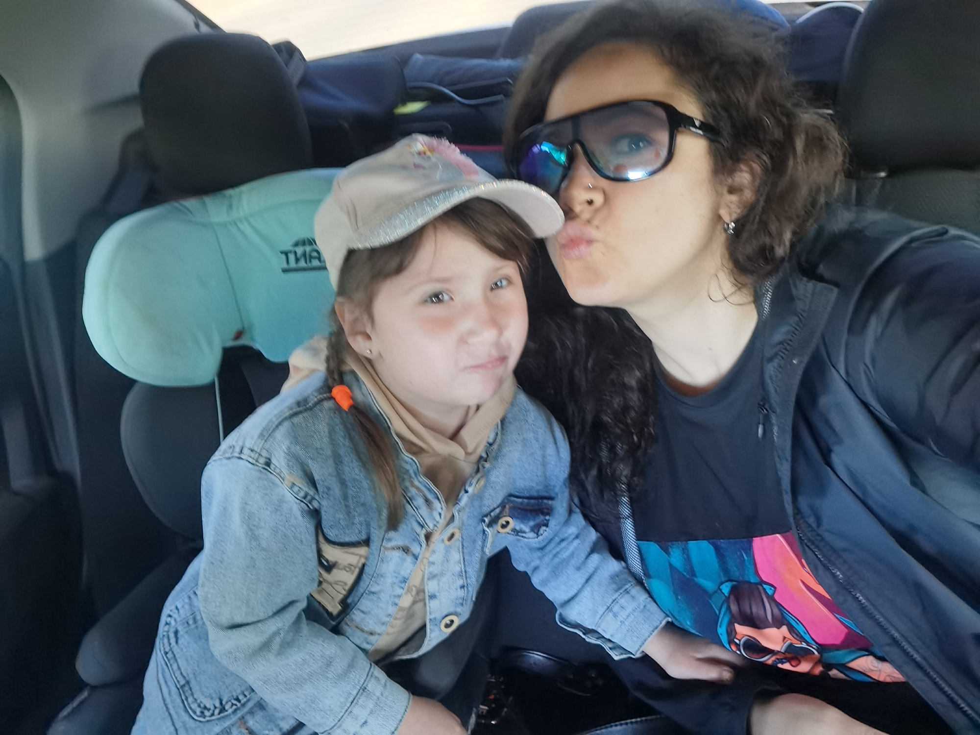 Two people, a woman and a young girl, are seated in the back of a car. The woman, on the right, is wearing sunglasses and making a kissing gesture towards the camera. She has curly dark hair and is wearing a dark jacket over a t-shirt with a colorful graphic. The young girl, on the left, is wearing a denim jacket over a tan hoodie and a baseball cap. She is looking towards the camera with a neutral expression. A light blue car seat cushion is visible behind the girl. The interior of the car is dark, with black seats and headrests. The lighting suggests it is daytime, likely outdoors. The image was taken in Chaikovsky, Russia.