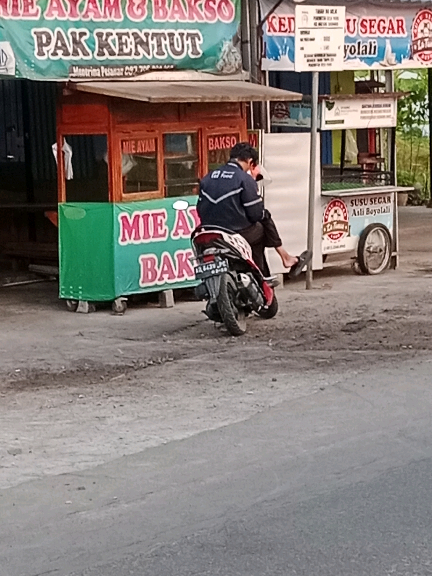 In Waru, Indonesia, a scene unfolds featuring a food stall and its patrons. A man wearing a dark jacket with white stripes is seated on a motorcycle, his arm around a woman who is also on the motorcycle. The food stall, identifiable by its green signage displaying "MIE AYAM BAKSO," is visible in the background. Another stall, this one advertising "SUSU SEGAR ASLI BOYOLALI," stands nearby. The environment appears to be a roadside setting, with a paved road in the foreground and a dirt or gravel area surrounding the stalls.