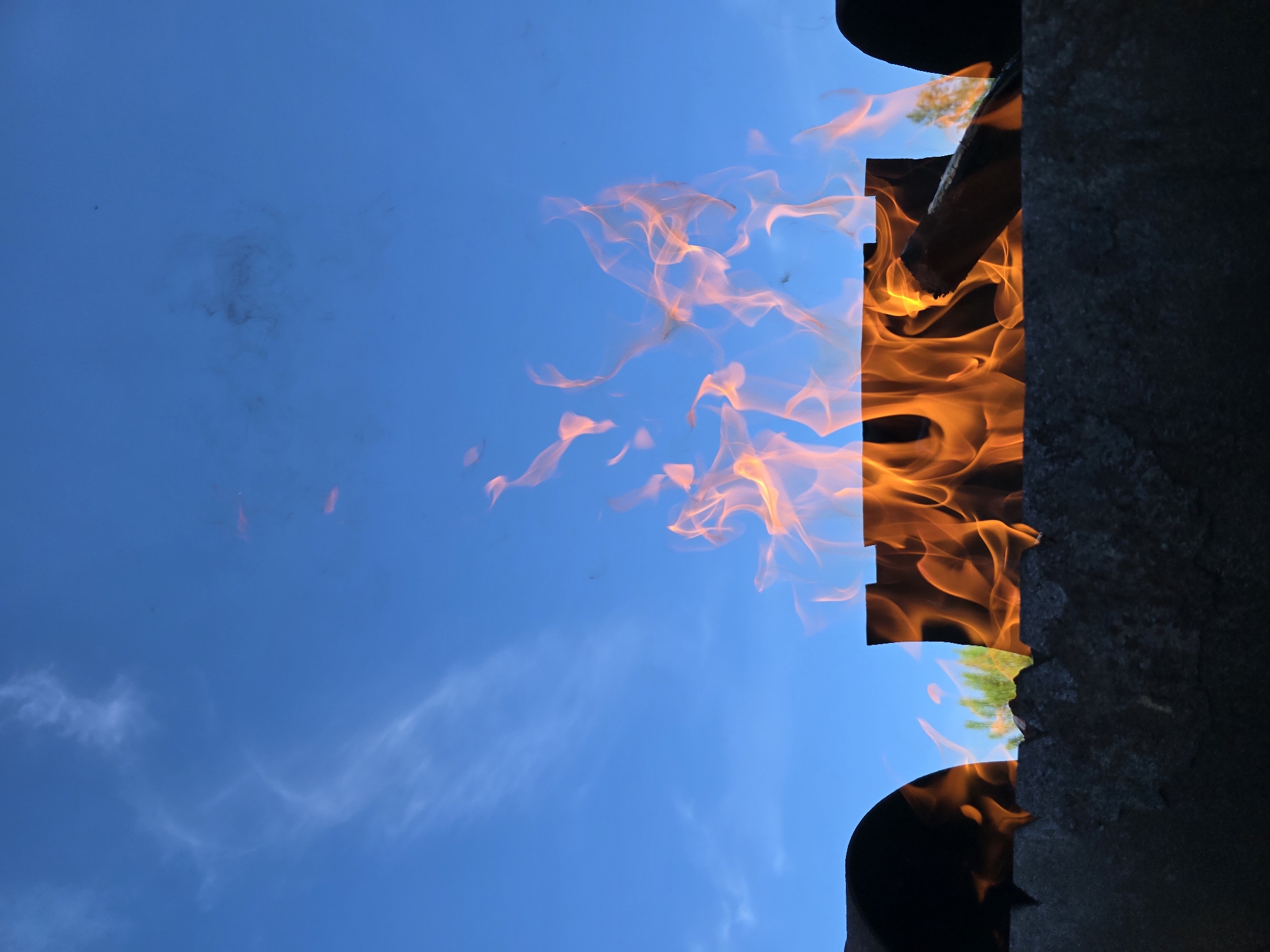 A close-up, low-angle shot depicts a fire burning in what appears to be a metal brazier or fire pit, set against a bright blue sky with wispy clouds. Flames, rendered in vibrant shades of orange and yellow, rise dramatically from the fire pit. The brazier itself is dark and textured, with the silhouette of a cylindrical object visible on the left and a similar object on the right, suggesting the fire pit may be enclosed or have raised sides. A piece of wood is visible within the flames. In the background, a blur of green foliage hints at trees. The scene is captured from a low perspective, emphasizing the fire and sky. The location is identified as Unknown, Unknown.
