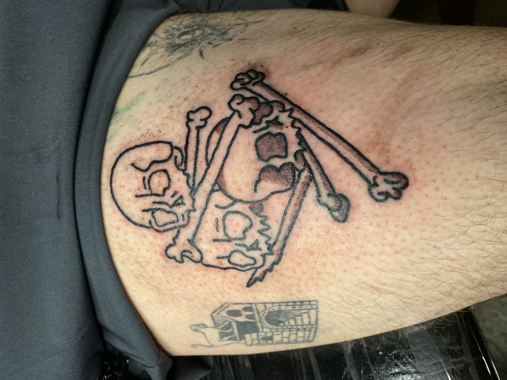 Close-up photograph of a person's upper arm/shoulder region, displaying two visible tattoos. The primary tattoo is an incomplete, cartoonish rendition of skulls and crossbones. Below this tattoo is another, smaller tattoo of a dark color. The person is wearing a dark gray or black shirt, the material of which is partially visible. The person's skin is fair, and there is visible hair. The background is out of focus and dark. No information about Ushuaia, Argentina is included in the picture.