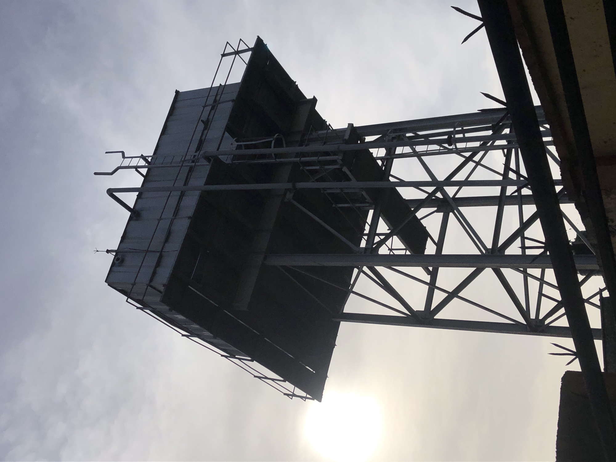 The image shows a low-angle view of a metal structure against a cloudy sky. The structure appears to be a large, possibly abandoned, billboard or sign support with a box-like section on top. The metal frame is visible, revealing a network of beams and supports. There are some rusted elements on the structure. The sun is visible through the clouds. Spiked elements are on top of the frame. The location is Unknown, Unknown.