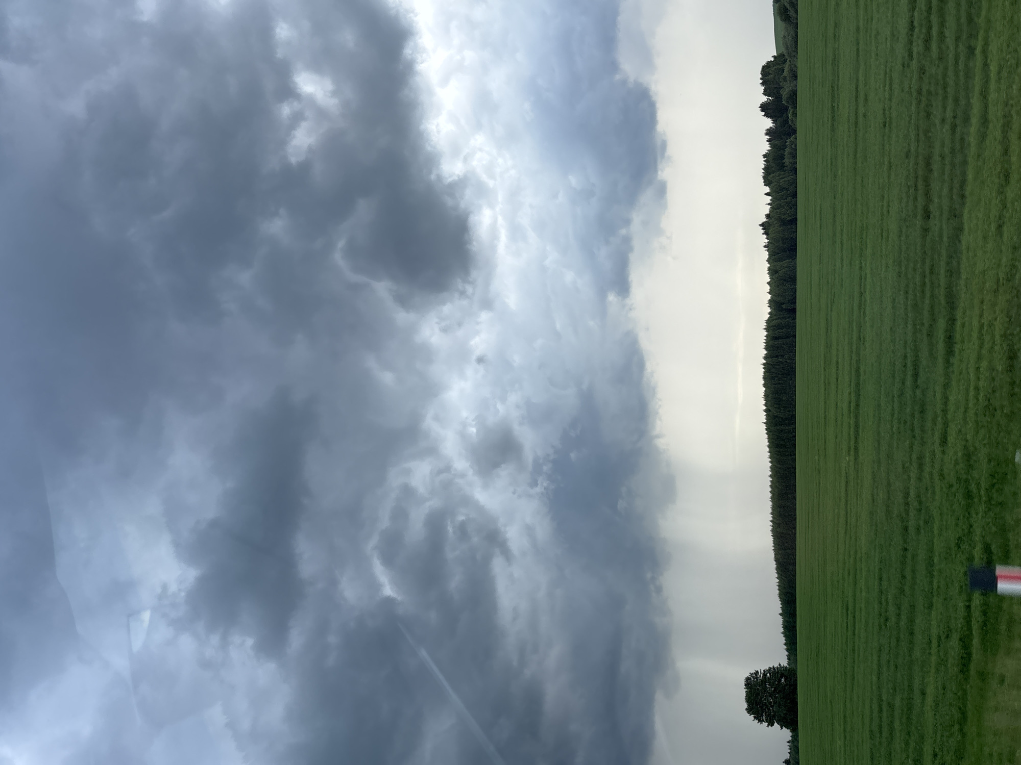 The image depicts a vast, green field under a dramatic, cloudy sky. In the distance, a dense line of dark green trees forms a horizon, punctuated by a solitary, large tree on the left side of the frame. Towards the center of the image, a curtain of rain is visible falling from the dark clouds, suggesting an approaching or ongoing storm. The foreground is dominated by the textured green field, with subtle horizontal lines indicating rows or growth patterns. A small portion of a red and white marker or post is visible at the bottom edge of the frame, on the right. The overall atmosphere is one of natural, somewhat somber beauty, characteristic of the rural landscape around Поварёнки, Russia.