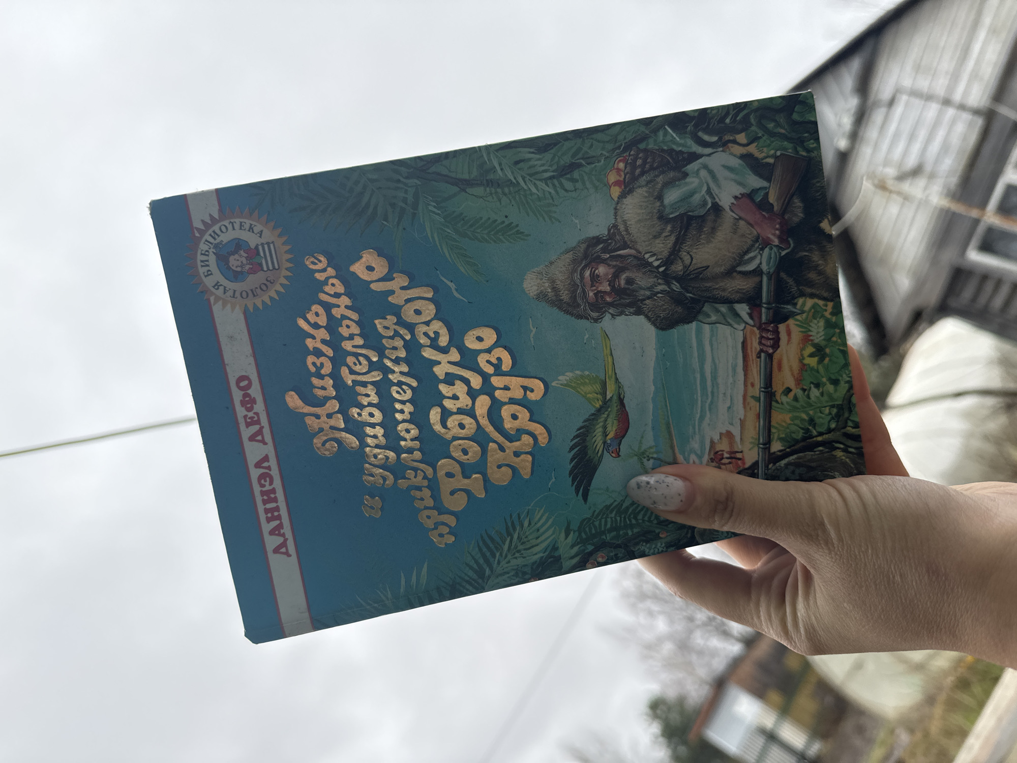 A woman is holding a book in her hand. The book is titled "Life and Adventures of Robinson Crusoe" in Russian and written by Daniel Defoe. The cover of the book is blue and features an illustration of Robinson Crusoe on a tropical island. The background is a blurry outdoor scene with a cloudy sky, a building, and some greenery.