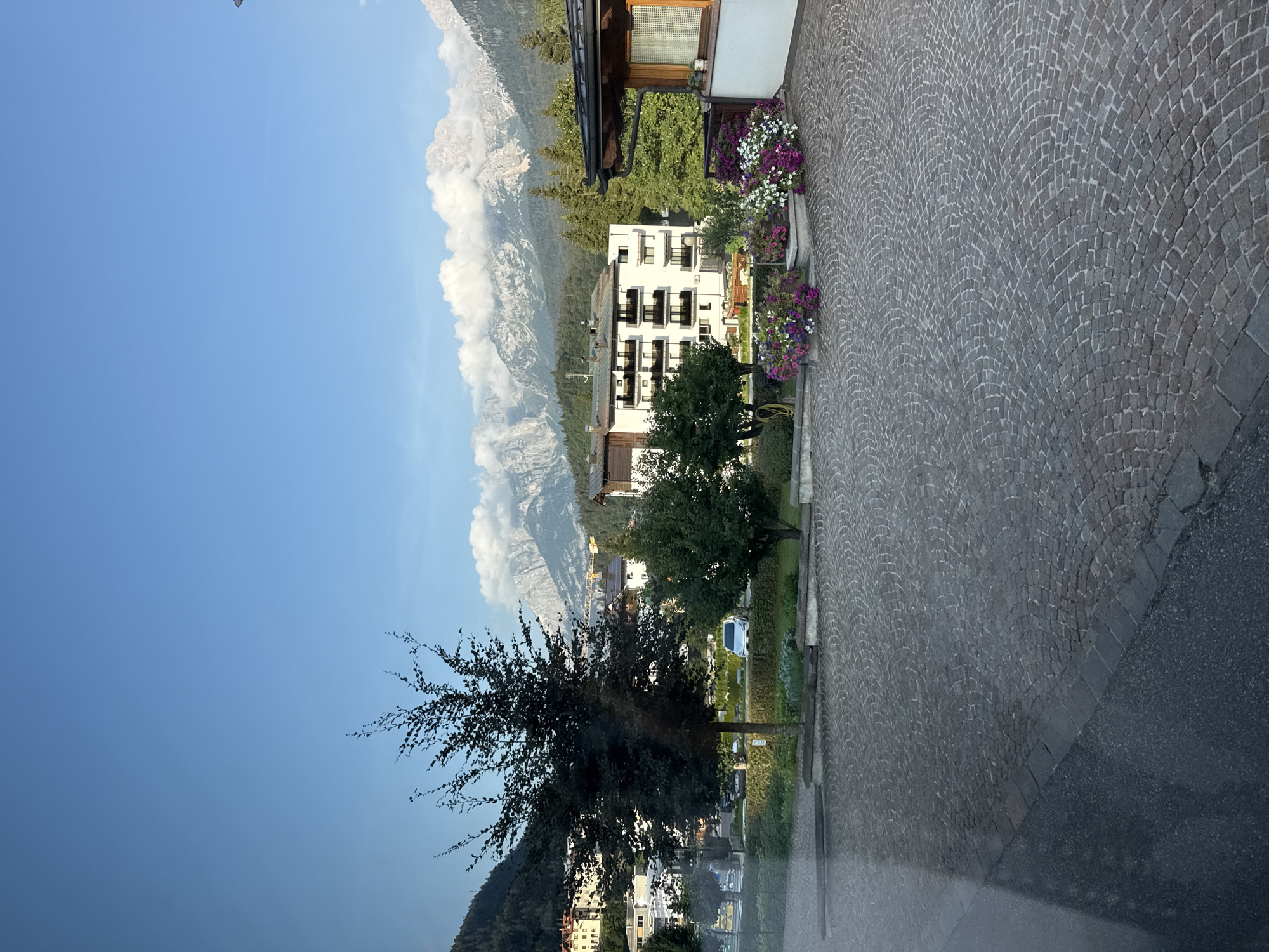 The image depicts a view from a paved area in front of a building, looking out towards a mountainous landscape. In the foreground, there is a cobblestone-paved area with a darker asphalt section to the left. Several planters filled with colorful flowers, including purple, pink, and white varieties, are arranged along the edge of the paved area and near a wooden-roofed structure.
In the middle ground, there is a residential building with multiple balconies and a light-colored facade. Trees and greenery are interspersed around the buildings, with a tall, dark-leaved tree prominent on the left side of the frame. A silver car is visible parked on the street in front of the building.
The background features a range of mountains, some with visible snow and cloud cover. The sky is a clear blue with scattered white clouds. A construction crane is visible in the distance to the right of the main building. The overall impression is that of a scenic town located in a mountainous region, likely during daylight hours. Based on the architecture and the mountainous setting, this scene is consistent with locations in the Dolomites, such as the town of Andalo, Italy. There are no people visible in the image.
