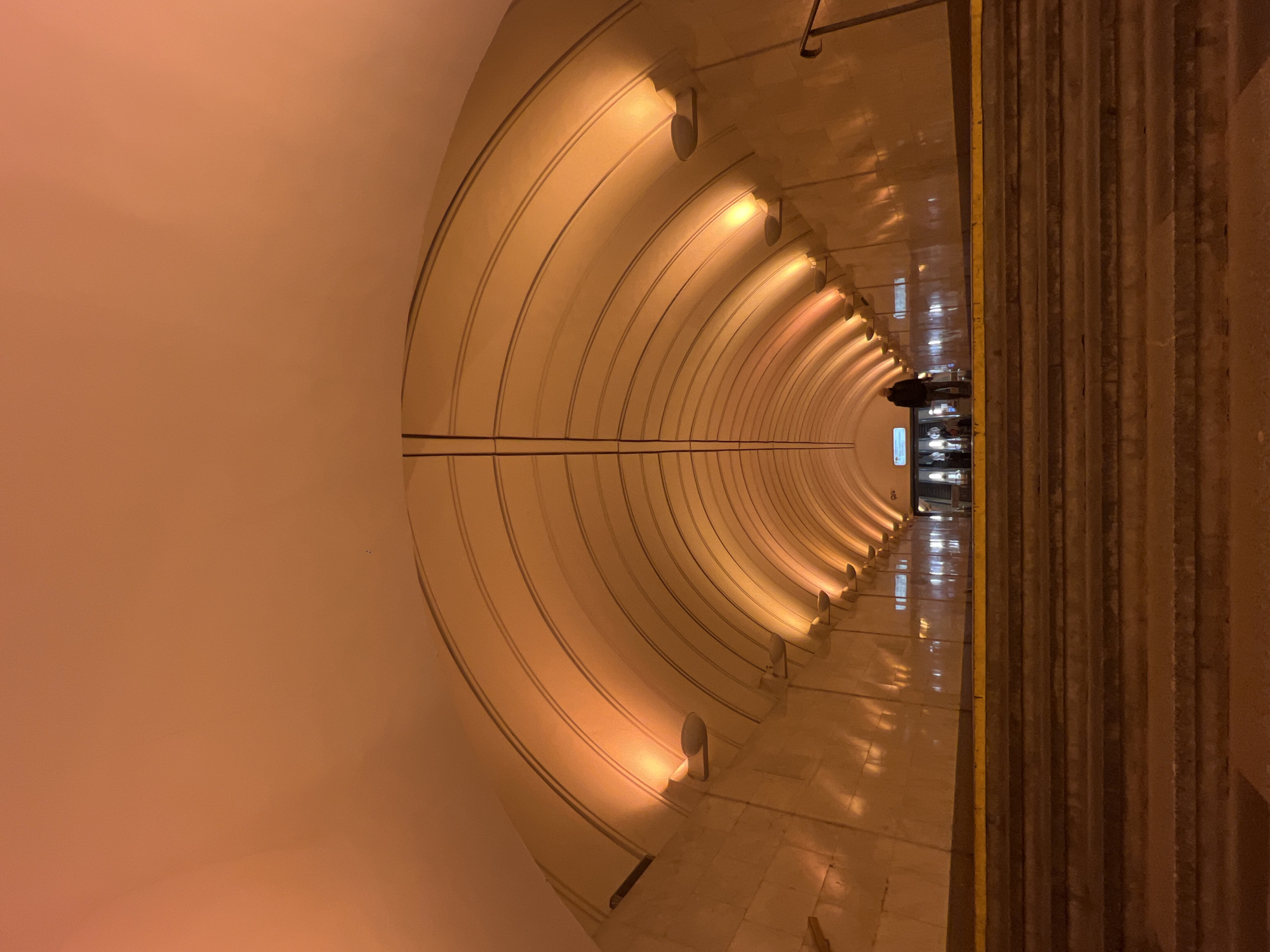 The image shows a brightly lit tunnel in a subway station, likely in Moscow, Russia, known for its ornate stations. The tunnel has a curved ceiling with evenly spaced lights illuminating the walls. The walls are lined with dark-colored stripes along the tunnel. The floor is tiled and reflective, with passengers standing at the end of the tunnel. To the right of the image is the side of a raised platform with stairs. The overall color scheme is warm, due to the lighting.