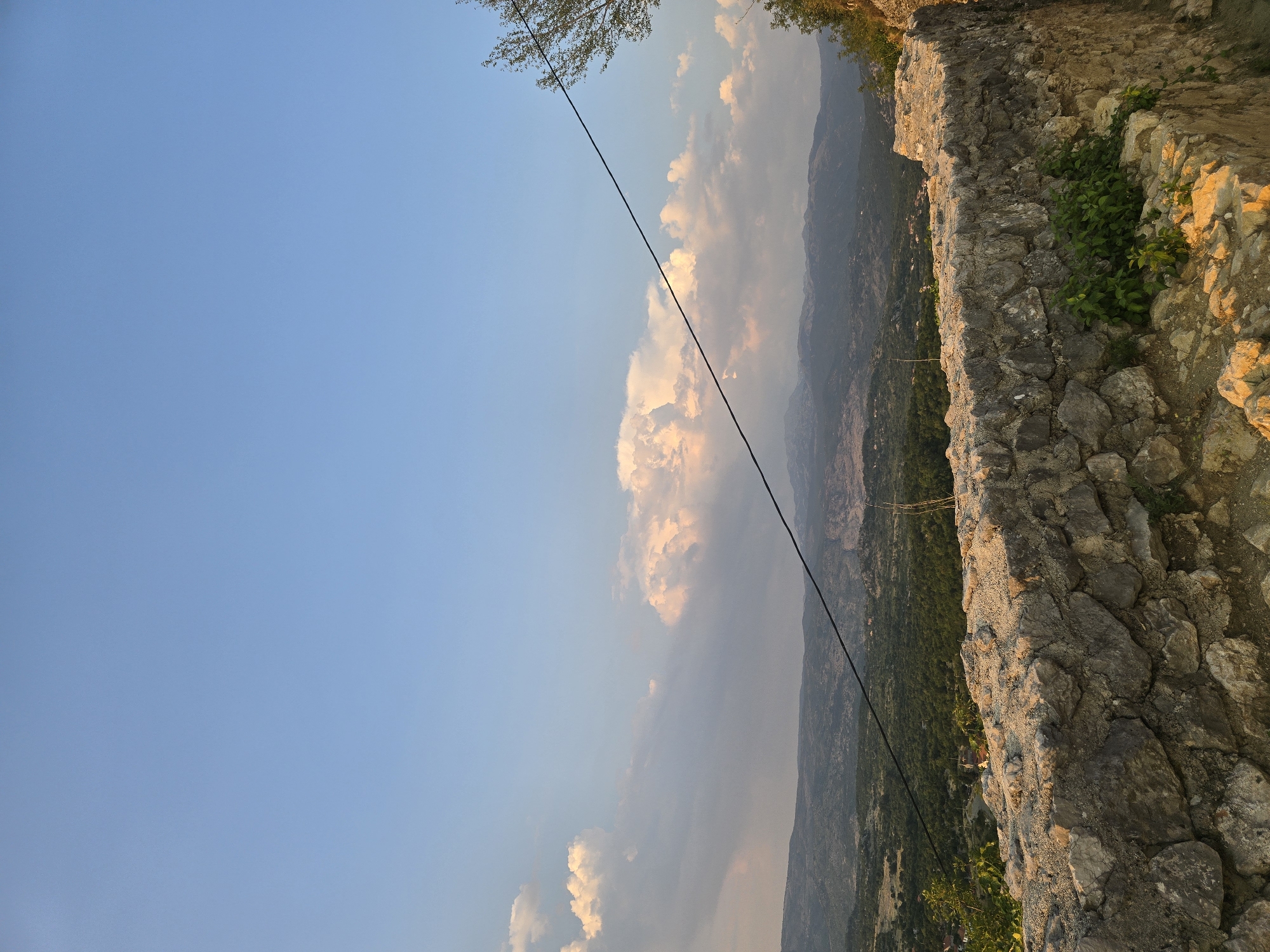 An outdoor, eye-level, wide shot captures a stone wall in the foreground of a mountain landscape under a clear blue sky. The stone wall, made of rough, weathered stones of varying sizes, forms a barrier that curves to the right. In some sections of the wall, green plants are growing between the stones. The landscape behind the wall is densely covered with green trees, with mountains visible in the distance. A black wire stretches diagonally across the frame from the top right to the middle left, cutting across the sky and the landscape. There are white and pinkish clouds clustered in the sky above the mountains. The sun is low on the horizon, casting a warm, golden light on the stone wall and parts of the landscape. The scene is devoid of any people or animals. The location is identified as Gornja Vrbica, Montenegro.