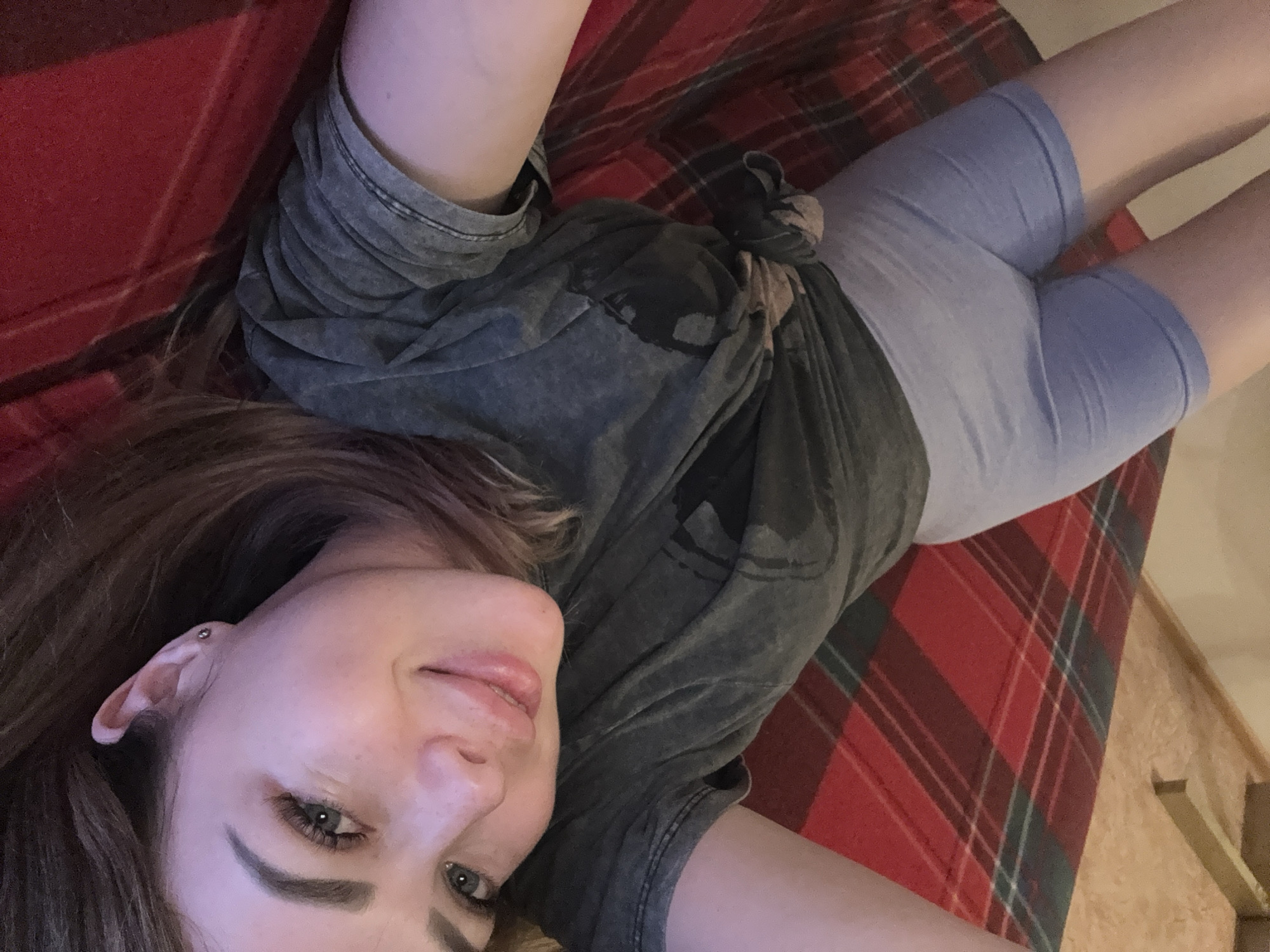 A young woman is lying on a red and green plaid blanket in an interior space. She is wearing a gray t-shirt and light blue shorts. Her face is turned towards the camera and she is smiling slightly. The city is unknown, as is the location.