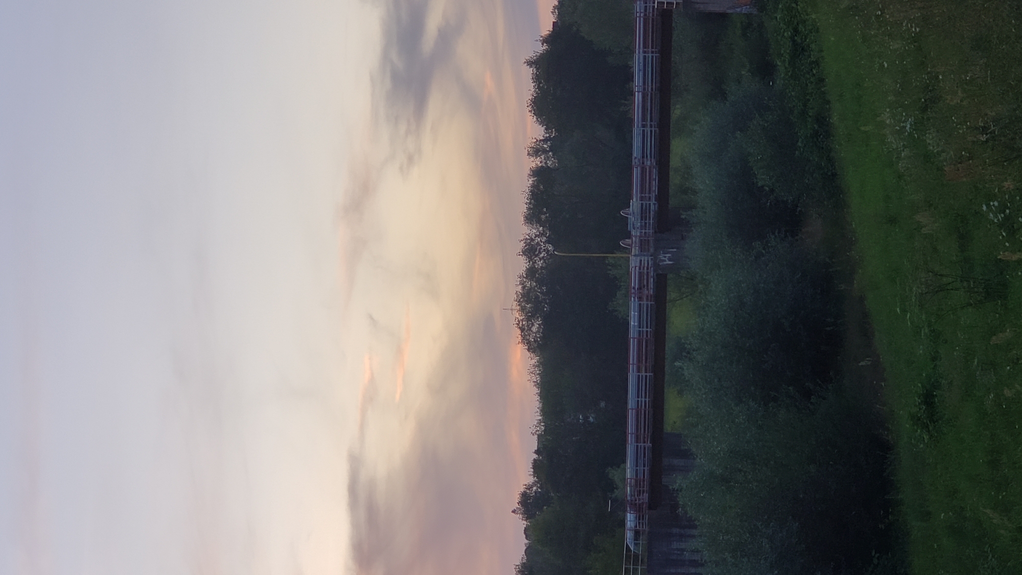 The image depicts a bridge over a body of water, possibly a river or stream, with lush greenery on either side. The sky above is a soft gradient of pale blue and pink, suggesting either sunrise or sunset. Trees line the banks of the water, and a street lamp is visible on the bridge. The bridge itself is a concrete structure with railings and appears to be supporting a road. The overall scene is serene and natural, with a hint of man-made infrastructure. The location is identified as Prešov, Slovakia.