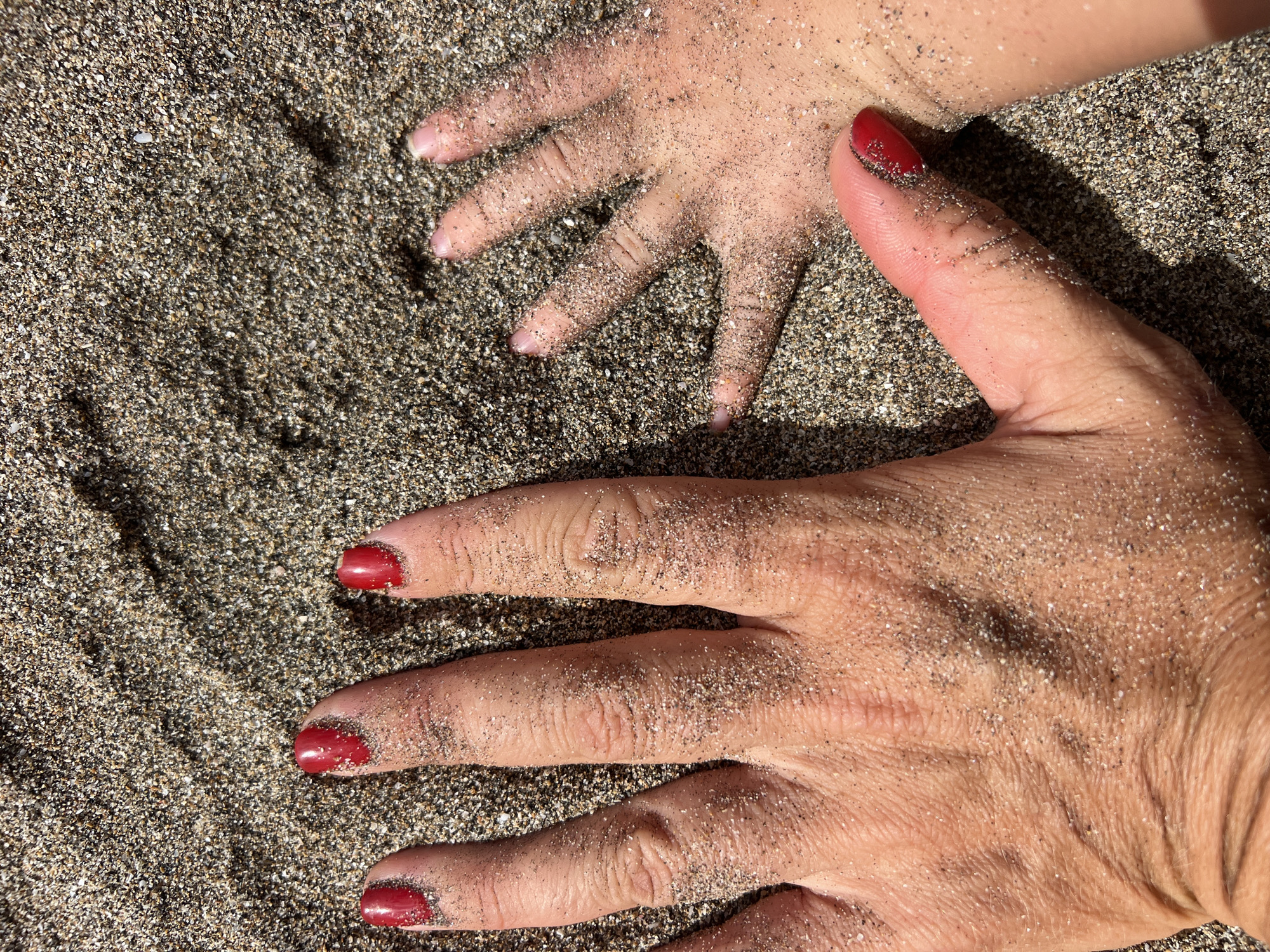 A close-up, high-angle shot shows two hands covered in sand on a beach in North Kuta, Indonesia. The left hand, presumably an adult's, is larger and positioned in the foreground, with fingers spread. Red nail polish is visible on its fingernails. The right hand, smaller and likely belonging to a child, is in the background, with fingers splayed. Both hands are partially obscured by the coarse, dark sand, which covers the entire frame. The lighting suggests it is daytime, with shadows cast by the hands and the uneven sand surface.