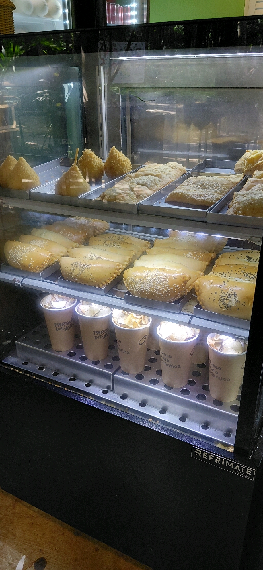 The image shows a refrigerated display case filled with various baked goods and snacks, likely in a cafe or bakery in Foz do Iguaçu, Brazil. The display case has two shelves. The top shelf contains what appear to be savory pastries, some coated in sesame seeds and others with a flaky, layered dough. There are also some cone-shaped fried snacks with a breadcrumb coating. The bottom shelf showcases more of the savory pastries, including some filled and folded over, also sprinkled with sesame or black seeds. In front of the pastries on the bottom shelf are several brown paper cups filled with what looks like potato chips or similar crispy snacks. The cups have some text and a logo printed on them. The background of the display case shows reflections of plants and possibly a bright green wall. A "REFRIMATE PREMIUM" label is visible on the front of the display case. The overall scene suggests a commercial establishment offering ready-to-eat food items.