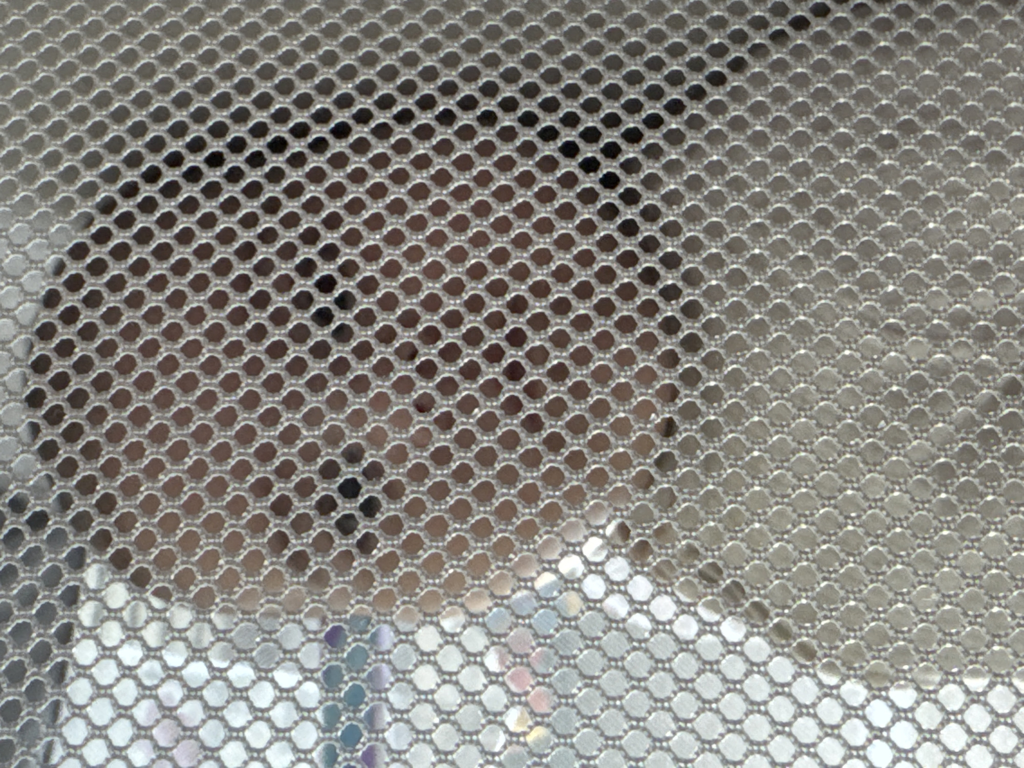 The image shows a close-up, abstract view of a person's head and shoulders seen through a silver mesh fabric. The mesh is a prominent foreground element, creating a patterned, honeycomb-like effect that obscures and distorts the subject behind it. The subject appears to have fair skin and is looking upwards or to the side. The lighting is bright, causing reflections on the mesh and highlighting parts of the subject's skin, creating a somewhat ethereal or diffused appearance. There are hints of other colors, possibly from clothing or the background, within the mesh openings, suggesting a colorful environment beyond the immediate subject. No specific location in Bălți, Moldova, can be identified from this image, as it is a close-up, abstract, and lacking any discernible environmental context.
