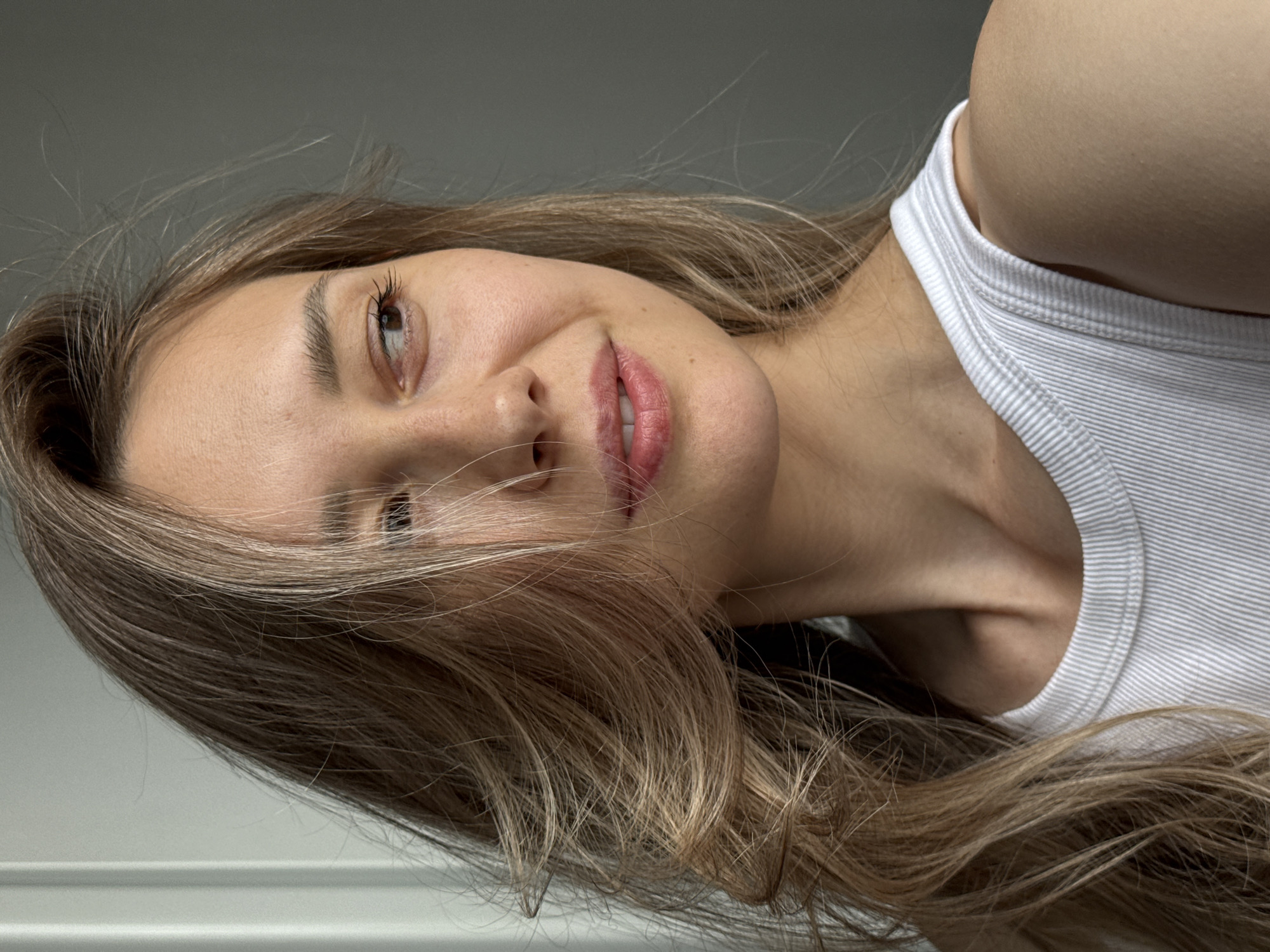 A woman with long brown and blonde hair lies on her side wearing a white tank top. She is smiling slightly and looking upwards. The background is a gray wall. There is no evidence of the location being Chaikovsky, Russia.
