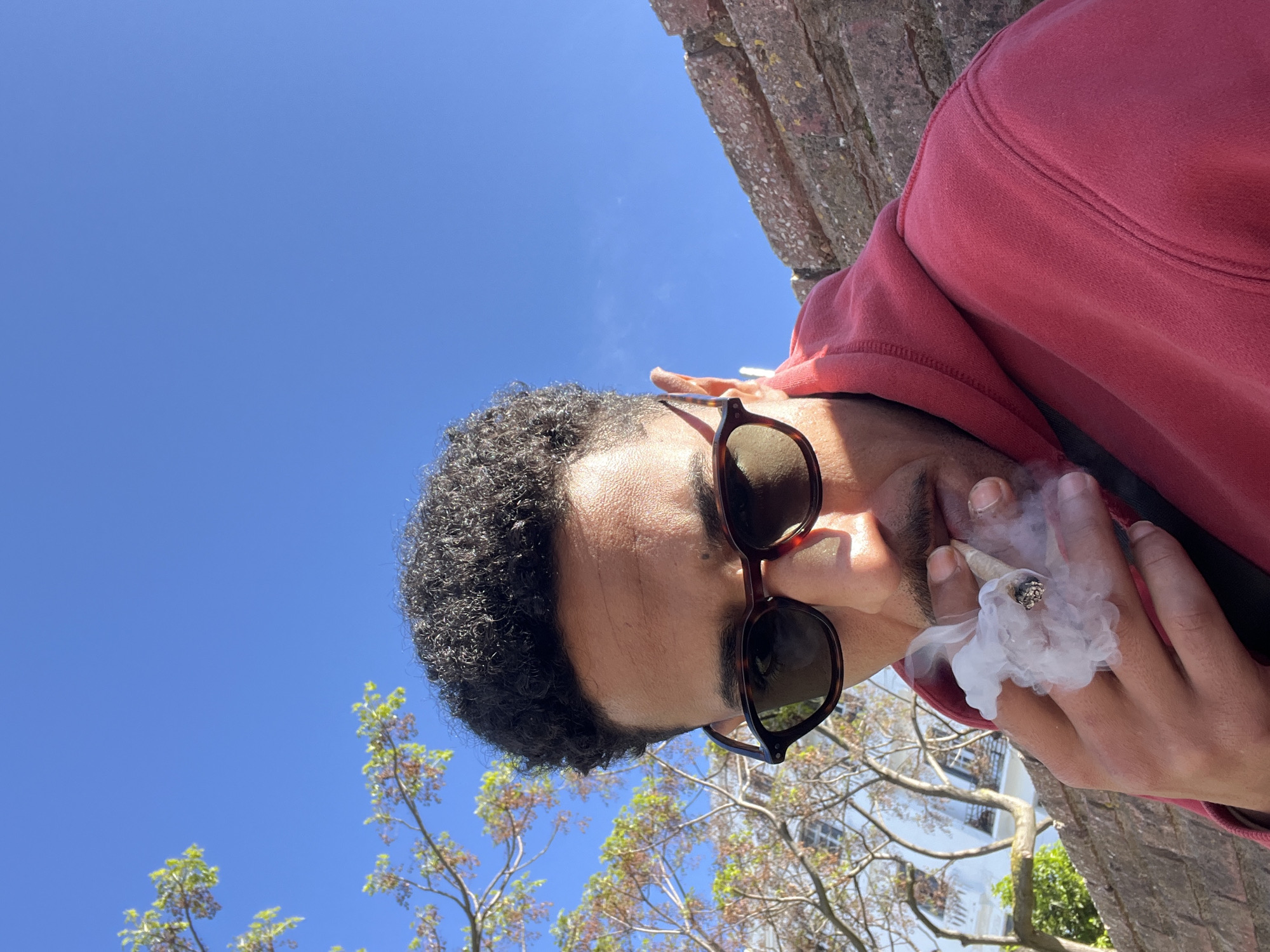 An upward-angle, outdoor image captures a male subject in Tanger, Morocco, positioned under a clear blue sky. The individual, with dark curly hair and a visible mustache, wears tortoise-shell framed sunglasses and a red hooded sweatshirt. He holds a lit, hand-rolled cigarette between his right thumb and forefinger, with a prominent plume of white smoke exhaling from his mouth area and curling around the cigarette's ember. In the background to the left, tree branches with green and some sparse reddish leaves are visible, partially obscuring a white building with dark-framed windows. To the right, behind the subject's shoulder, a reddish-brown brick wall forms part of the immediate environment.