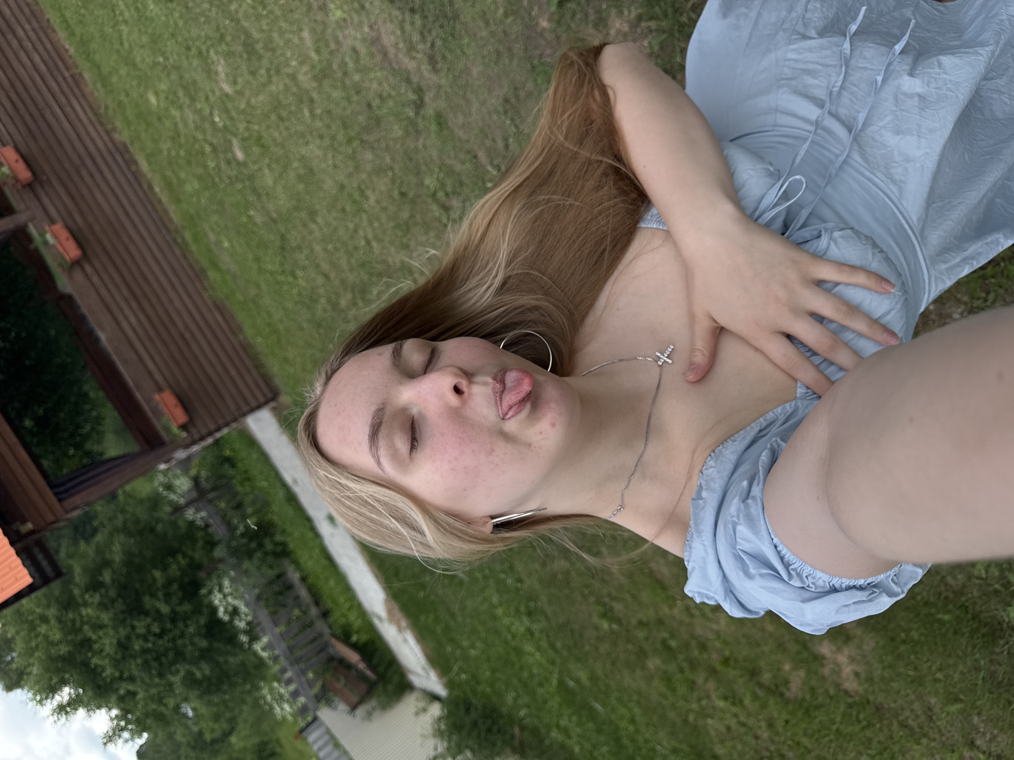 A young woman with blonde hair is taking a selfie outdoors, with her eyes closed and tongue sticking out. She is wearing a light blue off-the-shoulder dress and a silver cross necklace. Her right hand is placed on her chest. The background consists of a grassy area with a wooden fence and a building.