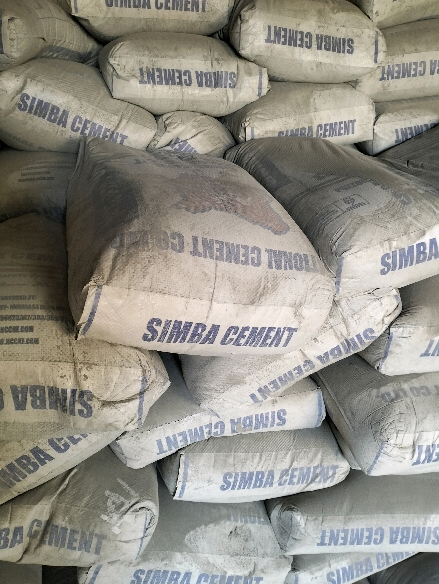 A high-angle, close-up shot captures a large stack of industrial sacks, primarily filled with cement, a common building material. The sacks are made of a woven, synthetic material, predominantly light gray and beige, with some showing signs of dust and wear. Prominently displayed on many of the sacks is the brand name "SIMBA CEMENT" in bold, dark blue lettering, suggesting a manufacturing or distribution hub in the Kinanie ward of Nairobi, Kenya, known for its industrial activities. The sacks are piled high, indicating a substantial quantity of stored product, likely awaiting transport or sale. The lighting is even, without harsh shadows, suggesting an indoor environment like a warehouse or storage facility. There are no people or animals visible in the frame, emphasizing the industrial and inanimate nature of the scene. The image conveys a sense of scale and the organized storage of bulk goods, characteristic of supply chain operations.
