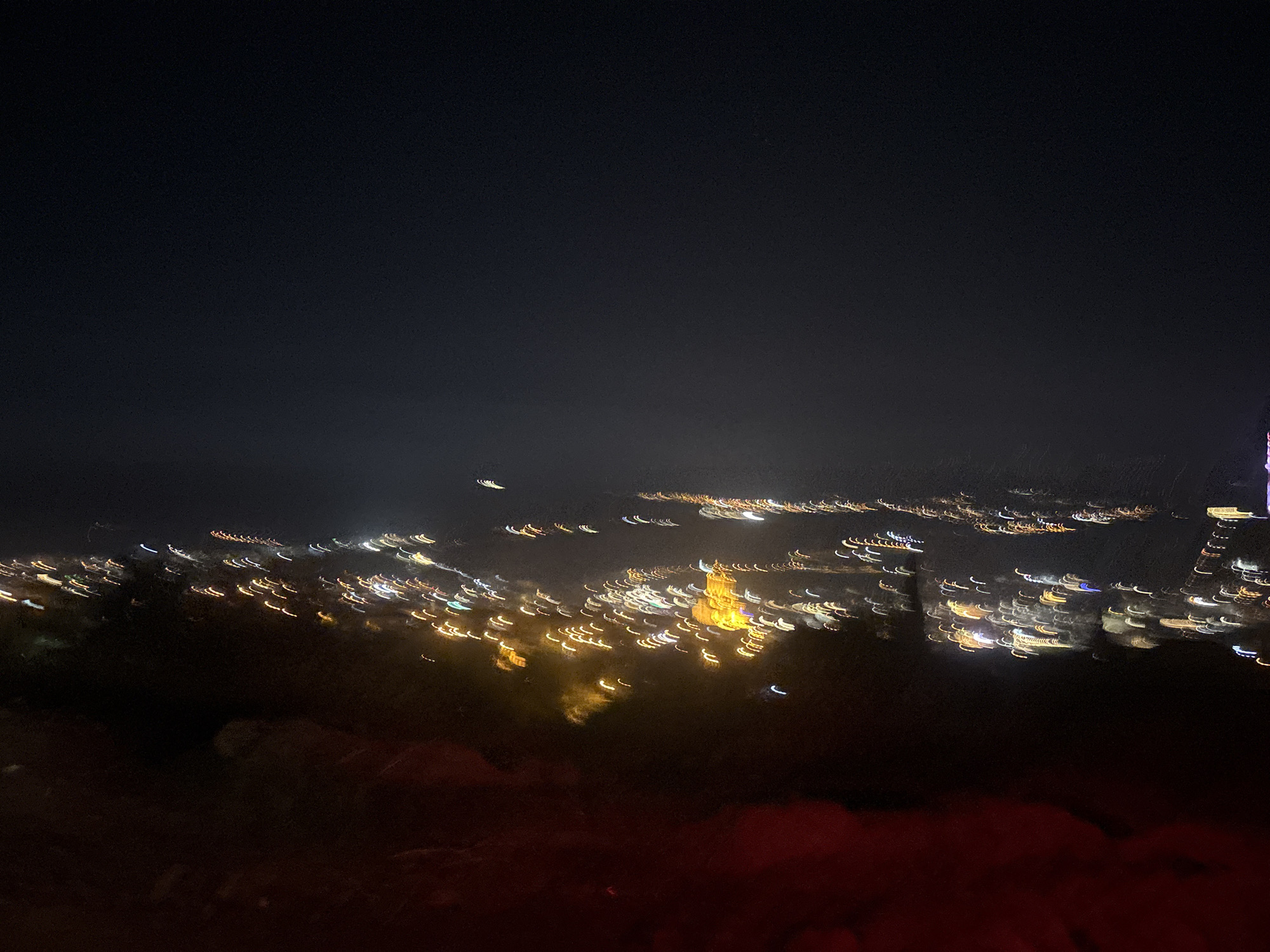 The image shows a blurred, long-exposure night view of the city of Tbilisi, Georgia. The focus point seems to be centered towards the center of the image where the lights look to be the brightest. In the background, there appears to be a large, dark sky. In the foreground, there are blurred objects that appear to be trees or other types of large landscape.