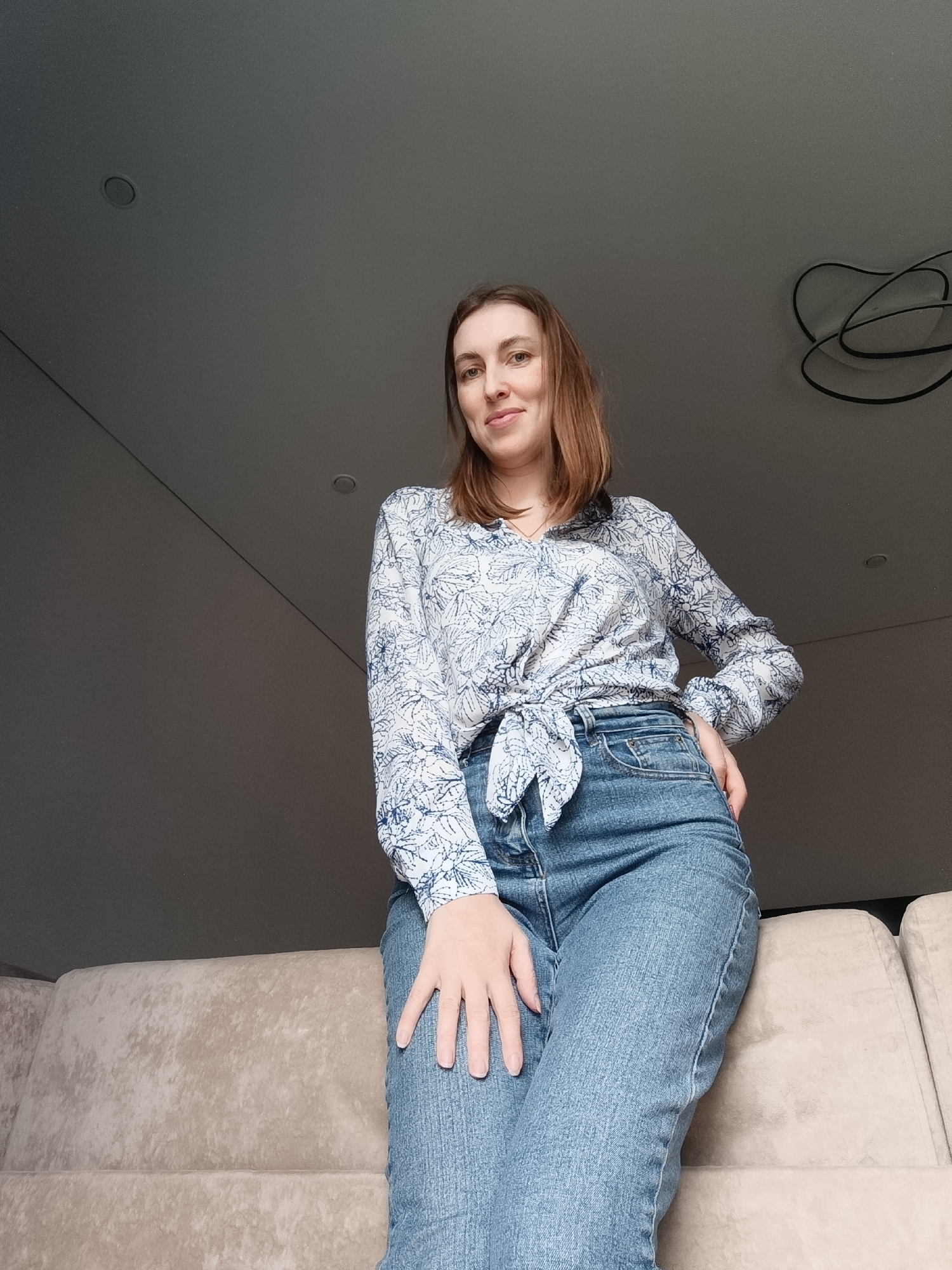 A woman is posing indoors. She is wearing a blue and white patterned shirt tied at the waist and blue jeans. Her left hand rests on her left leg. Her right hand is on her right hip. She is leaning against a beige couch. The ceiling is light grey and has two recessed lights and a black coiled wire fixture. The location is Chaikovsky, Russia.