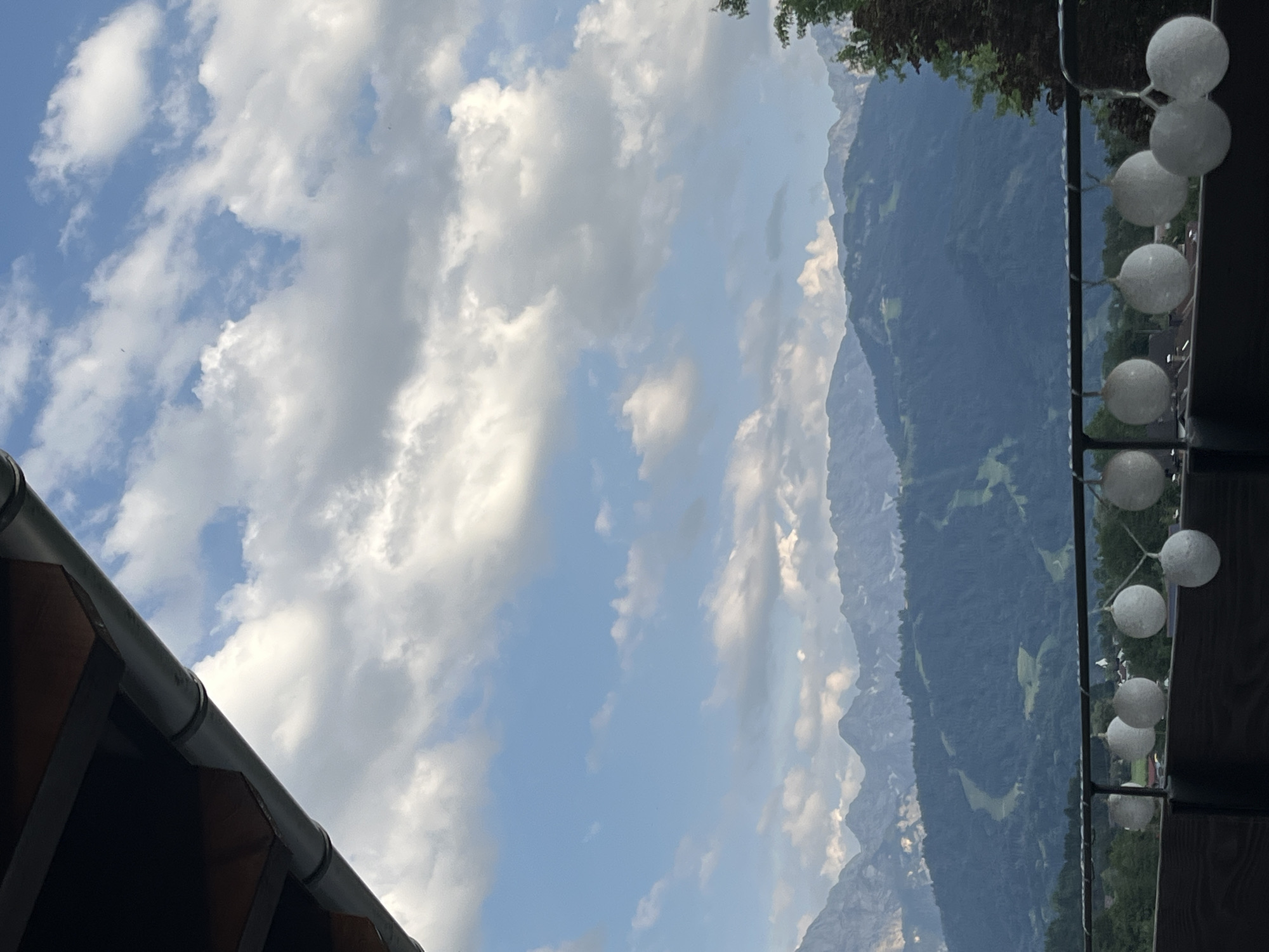 The image captures a scenic view of the Bavarian Alps from an elevated perspective, likely a balcony or terrace. The scene is dominated by a vast expanse of blue sky filled with dramatic, white, fluffy clouds. To the right, the rugged, dark blue slopes of the mountains rise sharply, partially obscured by the evening sky. Lush green trees line the upper slopes, hinting at the verdant alpine landscape.
In the foreground, on the right side of the image, a string of white, spherical lights hangs from a railing, suggesting an outdoor seating area or a decorated venue. These lights, illuminated against the darkening sky and mountain backdrop, add a touch of ambiance. On the left, a section of a dark-colored roof and gutter system is visible, framing the scene and implying the presence of a building.
The overall impression is one of serene natural beauty, with a touch of man-made comfort provided by the string lights. The time of day appears to be late afternoon or early evening, given the subdued lighting on the mountains and the presence of the decorative lights. The weather seems pleasant, with a mix of sun and clouds. The location cues, such as the distinct mountain range and the implied European setting, align with Garmisch-Partenkirchen, Germany. There is no visible text in the image.