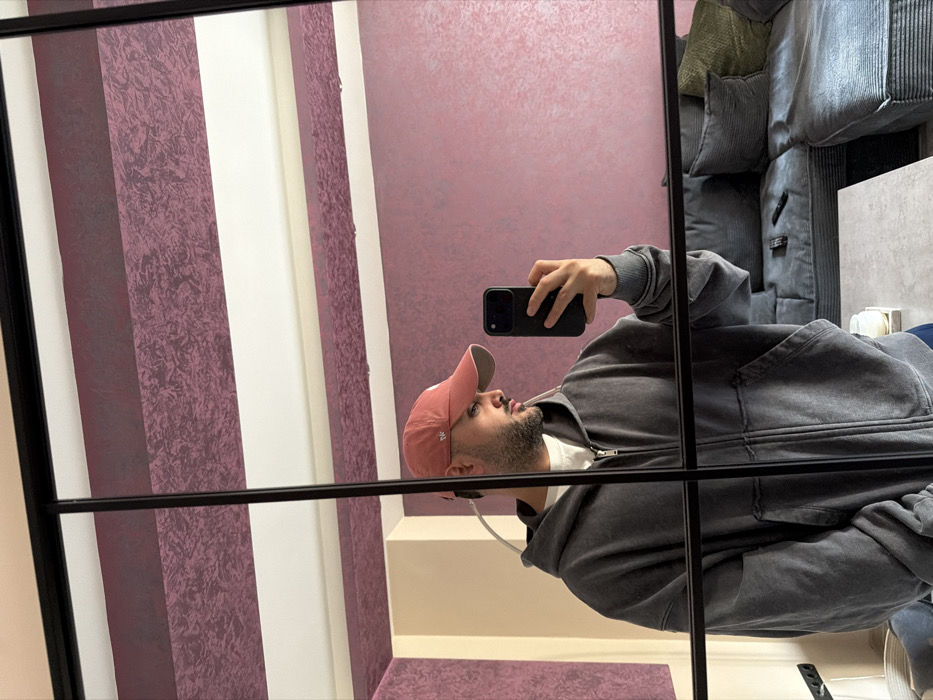 A male individual, estimated to be in his mid-20s to early 30s, is depicted in a mirror selfie within an interior setting, assumed to be located in Sofia, Bulgaria. He has short dark hair visible beneath a faded reddish-pink baseball cap worn backwards, and a short beard or heavy stubble. He is wearing an oversized, charcoal grey, washed-out zip-up hoodie over a white crew-neck t-shirt. His right hand holds a dark-colored smartphone, equipped with a prominent camera module, pointed towards the mirror to capture the image. His head is turned slightly to his right, away from the phone's lens.
The background, visible in the mirror's reflection, reveals a room with distinctive wall treatment: broad horizontal stripes of textured, mottled deep maroon or plum alternate with wider sections of plain off-white or light beige. A large, grey sectional sofa, upholstered in a ribbed or corduroy fabric and adorned with multiple cushions, occupies the right side of the reflected space. A black television remote control is positioned on the sofa. A light-colored, rectangular coffee table is present in the foreground of the reflected image, with a white decorative object or candle visible on its surface. The overall image is framed and sectioned by the black bars of a multi-paneled mirror. No specific street or building within Sofia is identifiable from the provided visual information.