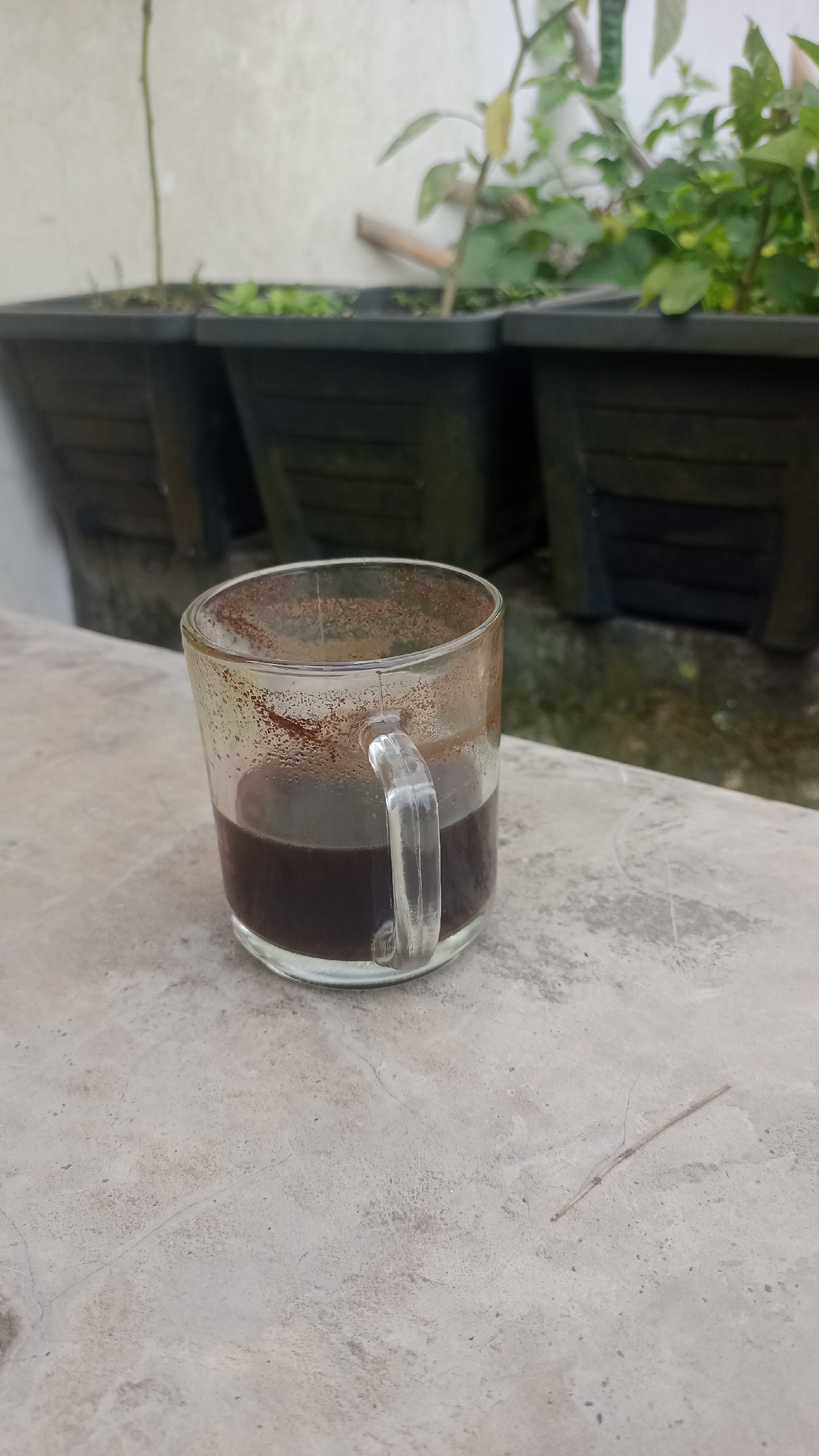 A clear glass mug containing dark liquid, likely coffee, rests on a light-colored, textured surface, possibly concrete. The mug has a thick, curved handle. The rim of the mug is dusted with fine brown powder, suggesting coffee grounds. In the background, several black plastic planters are visible, each containing green plants. The planters are arranged in a line against a textured wall. The overall setting appears to be outdoors, possibly on a patio or balcony, in Pakemitan, Indonesia. The scene is static and unpopulated.
