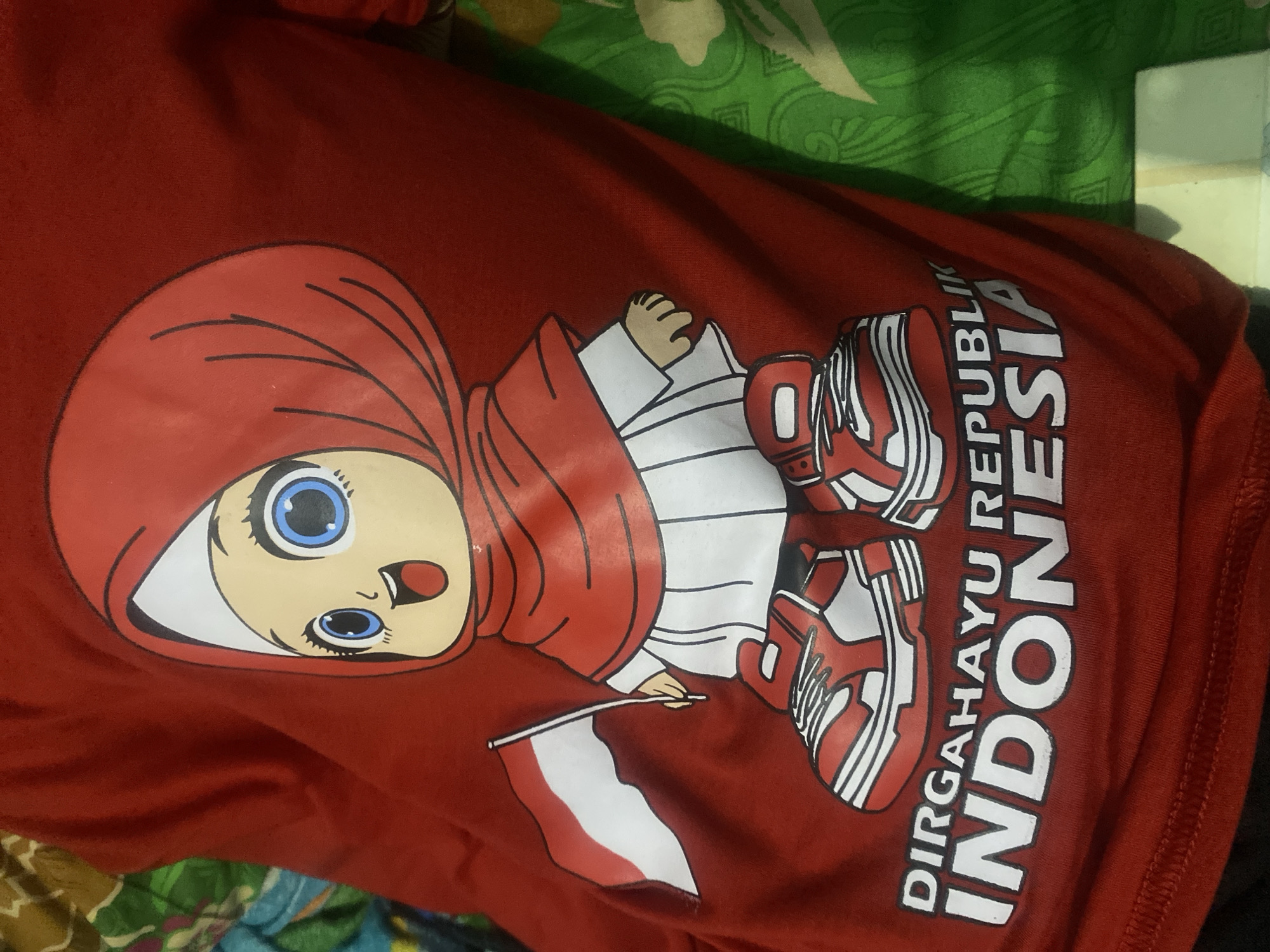 The image shows a red t-shirt with a cartoon illustration of a young girl wearing a red hijab and holding the Indonesian flag. The girl has large blue eyes and a cheerful expression. She is wearing a white shirt and skirt, and red and white sneakers. Below the illustration, text reads "DIRGAHAYU REPUBLIK INDONESIA" in white letters. The t-shirt is likely being displayed or worn, as evidenced by the folds and creases in the fabric. The background is blurred and appears to be a patterned fabric, possibly a bedsheet or curtain, in shades of green and brown. The image quality is adequate, though some areas are slightly out of focus. The scene is indoors. This image is a photograph of a t-shirt, not a depiction of a specific location in Kalitinggar, Indonesia.