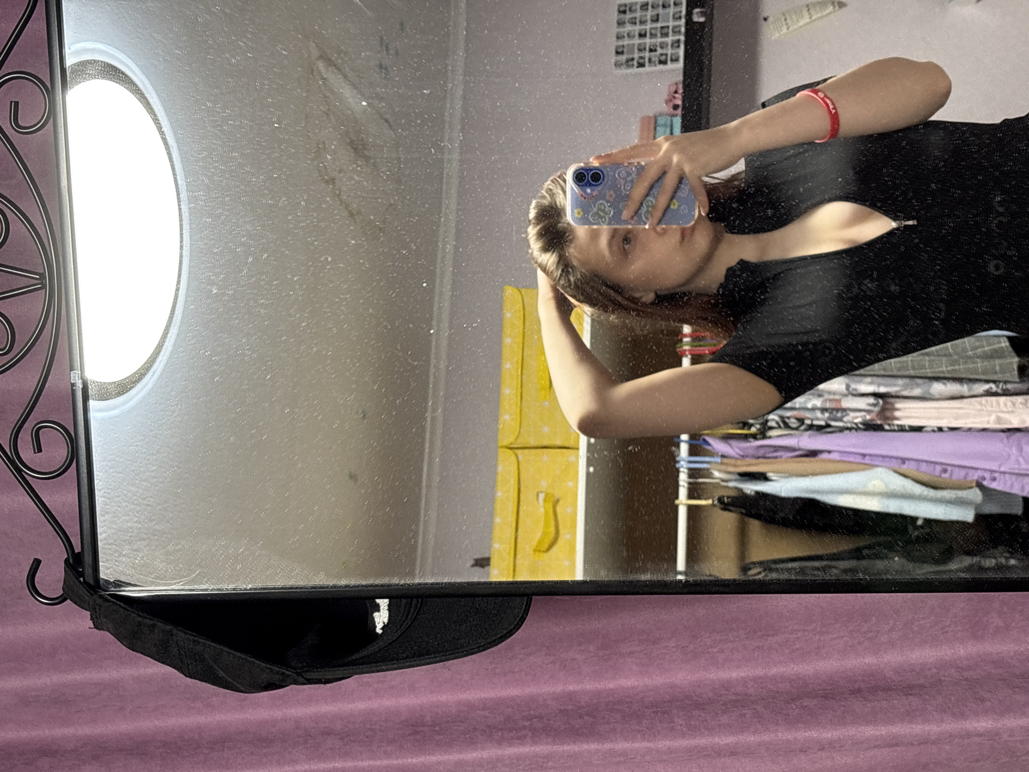 A person is taking a selfie in a mirror. The reflection shows the person wearing a black top and holding a phone with a blue case. A red bracelet is visible on the person's wrist. The background includes a closet with clothes, a yellow box, and a wall with pictures. A black cap is placed on the bottom of the mirror. The photo appears to be taken indoors. The location in the city of Прикамский, Russia is not discernible from the image.