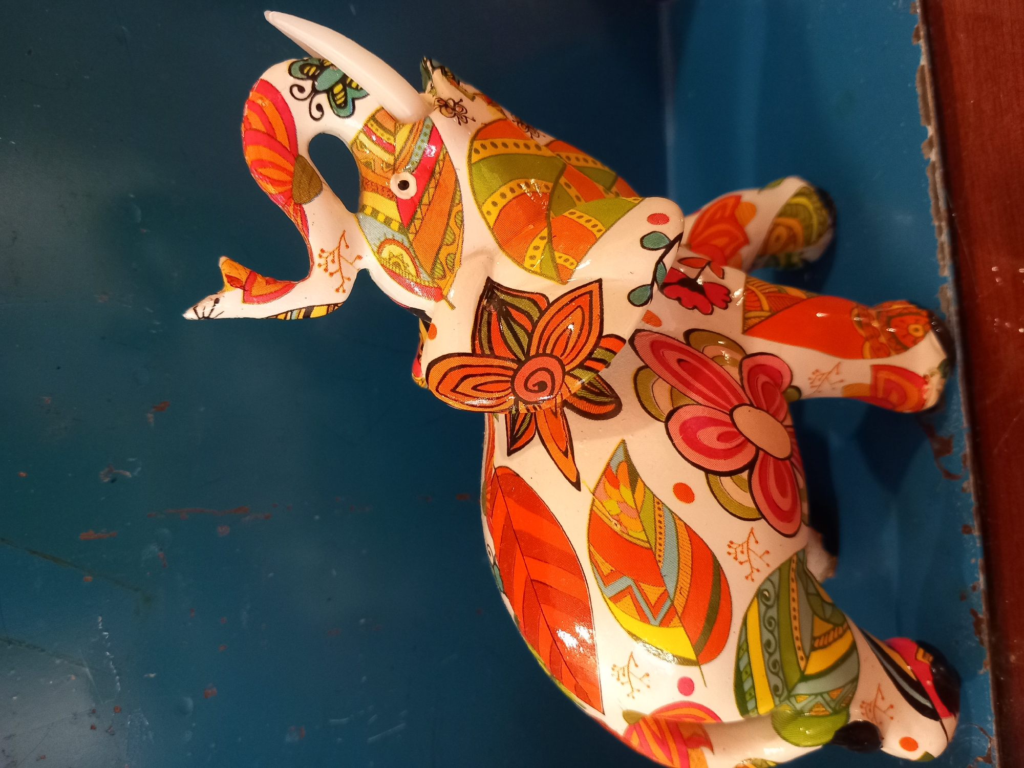 A white ceramic elephant figurine, adorned with vibrant floral and leaf patterns in shades of orange, yellow, green, and pink, stands on a blue surface. The elephant's trunk is raised, and one tusk is visible. The background is a solid, dark blue. The scene is an indoor, unknown location.