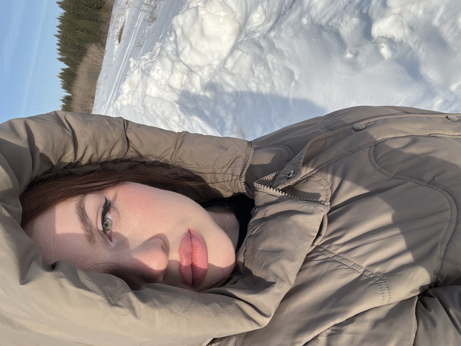 The image depicts a young woman in a snowy outdoor environment, identified as Pavlovka, Russia. The woman is positioned from the chest up, angled slightly to the viewer's left, with her gaze directed at the camera. She has reddish-brown hair, visible around her face, green eyes accentuated with dark eyeliner, and full lips colored with pink lipstick. She is wearing a light brown or taupe quilted puffer jacket with the hood drawn up, partially shadowing the left side of her face. A black collar or garment is visible beneath the jacket's opening. Strong natural light illuminates the scene, creating shadows on her face and clothing. The background consists of a vast expanse of white snow, showing some irregularities and potential tracks. To the right, the snow is visibly mounded. In the mid-ground, a line of leafless trees is visible, followed by a dense coniferous forest under a clear, bright blue sky.
