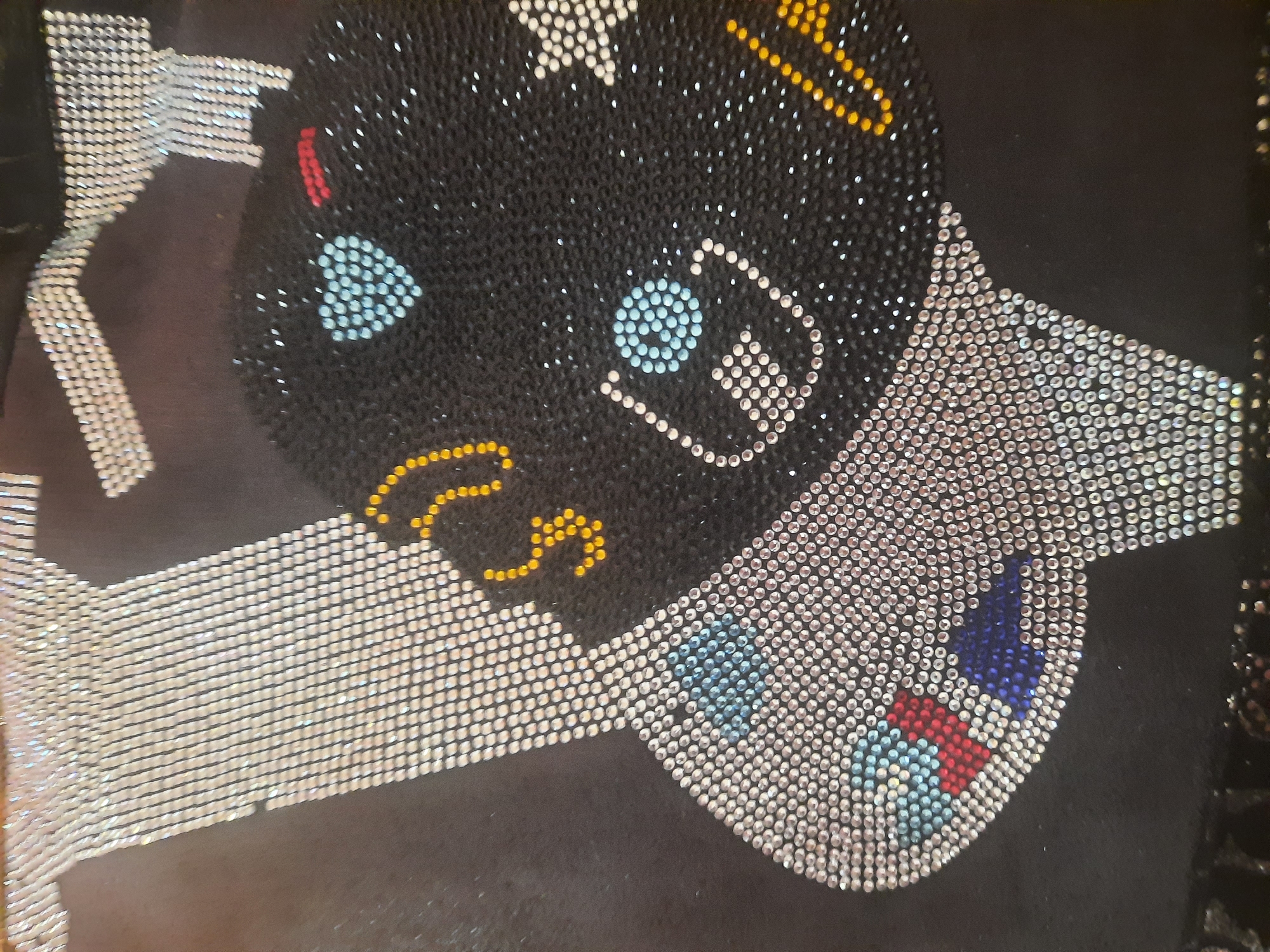 The image is a close-up, angled shot of a black fabric adorned with rhinestones. The rhinestones are arranged to form a design that appears to be a stylized depiction of a face or head. The design features a large, black, rounded shape representing the head, outlined and partially filled with rhinestones. There are various colored rhinestones used for details, including a light blue heart on the cheek, yellow circular shapes, red dots, and a white star shape. The face is angled towards the left of the frame, with some of the design extending upwards and to the left, appearing as a white, elongated shape with a textured surface formed by rhinestones. There is no discernible location in the city of Unknown, Unknown.