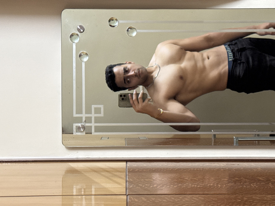 A man is captured in a mirror selfie, shirtless and posing to display his physique. He is leaning or reclining, with his body angled across the frame. He holds a light-colored smartphone with a triple camera setup in his right hand, reflecting his face and upper body. He wears dark pants or underwear with a visible blue waistband that has "HUGO BOSS" written in white lettering. Around his neck is a silver chain, and on his right wrist, he wears a gold-colored bracelet. His expression is neutral to slightly serious.
The mirror itself is rectangular and features decorative etched patterns, including geometric lines forming squares and other shapes, along its borders. Several clear, circular crystal or glass embellishments are embedded along the top and left edges of the mirror. The background visible through the mirror is a plain, light-colored wall.
Below the mirror, there is a piece of furniture, likely a dresser or cabinet, made of wood. The left section of the furniture has a glossy, light wooden finish, while the right section appears to have a darker, more textured wood grain. The surfaces reflect light and objects. The overall setting appears to be indoors, well-lit, possibly a bedroom or dressing area. The image is taken in Kolkata, India.