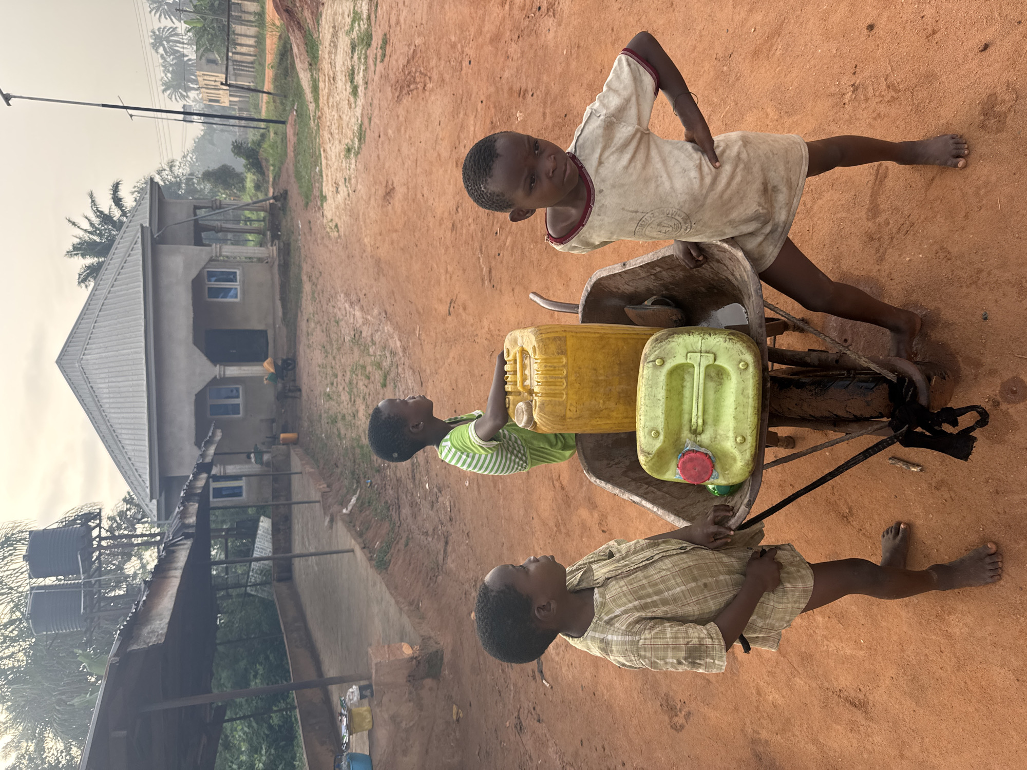 Three barefoot children are near a wheelbarrow with two yellow and green plastic containers. They are on a red dirt road or yard in Eziudo, Nigeria. In the background is a building with a gray roof, and the sky is overcast.