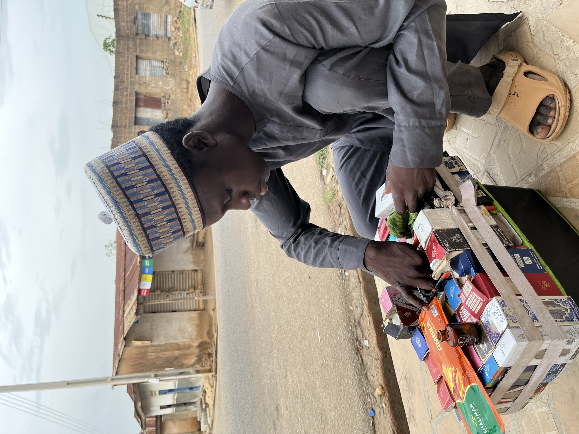 A young man wearing a traditional Hausa cap and gray long-sleeved shirt is kneeling next to a collection of items secured by straps, possibly for sale. The items include packaged incense sticks and other small boxes. He is wearing tan-colored sandals. He appears to be arranging or selecting an item from the collection.
The scene is outdoors, likely a street in Jos, Nigeria, suggested by the architecture of the buildings in the background, which feature weathered facades and corrugated metal roofing. The street surface is asphalt. The setting appears to be a modest urban environment.