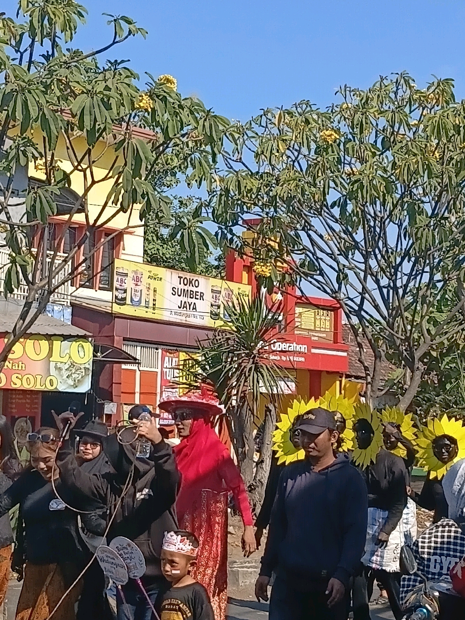In Wadungasri, Indonesia, a street scene unfolds under a clear blue sky with abundant sunshine illuminating a procession. Several individuals, some adorned with sunflower costumes, participate in what appears to be a cultural event or parade. In the foreground, a young boy in a black t-shirt with a white and red headdress stands looking to the right. Behind him, a man in a dark blue hoodie and black baseball cap walks forward, while several people in black clothing and sunflower headpieces are also visible.
To the left, a woman wearing a black hijab and sunglasses holds a microphone and appears to be speaking. Beside her, another woman in a black hijab and sunglasses holds a bottle of water. A third woman to their right wears a bright red hijab and a long red and gold patterned skirt, also holding a microphone. In the background, a yellow building with red accents displays signage for "Toko Sumber Jaya" and "ABC POWER." Trees with green leaves and yellow flowers are scattered throughout the scene, adding to the vibrant atmosphere. The presence of microphones and the festive attire suggest a public event with a celebratory nature.