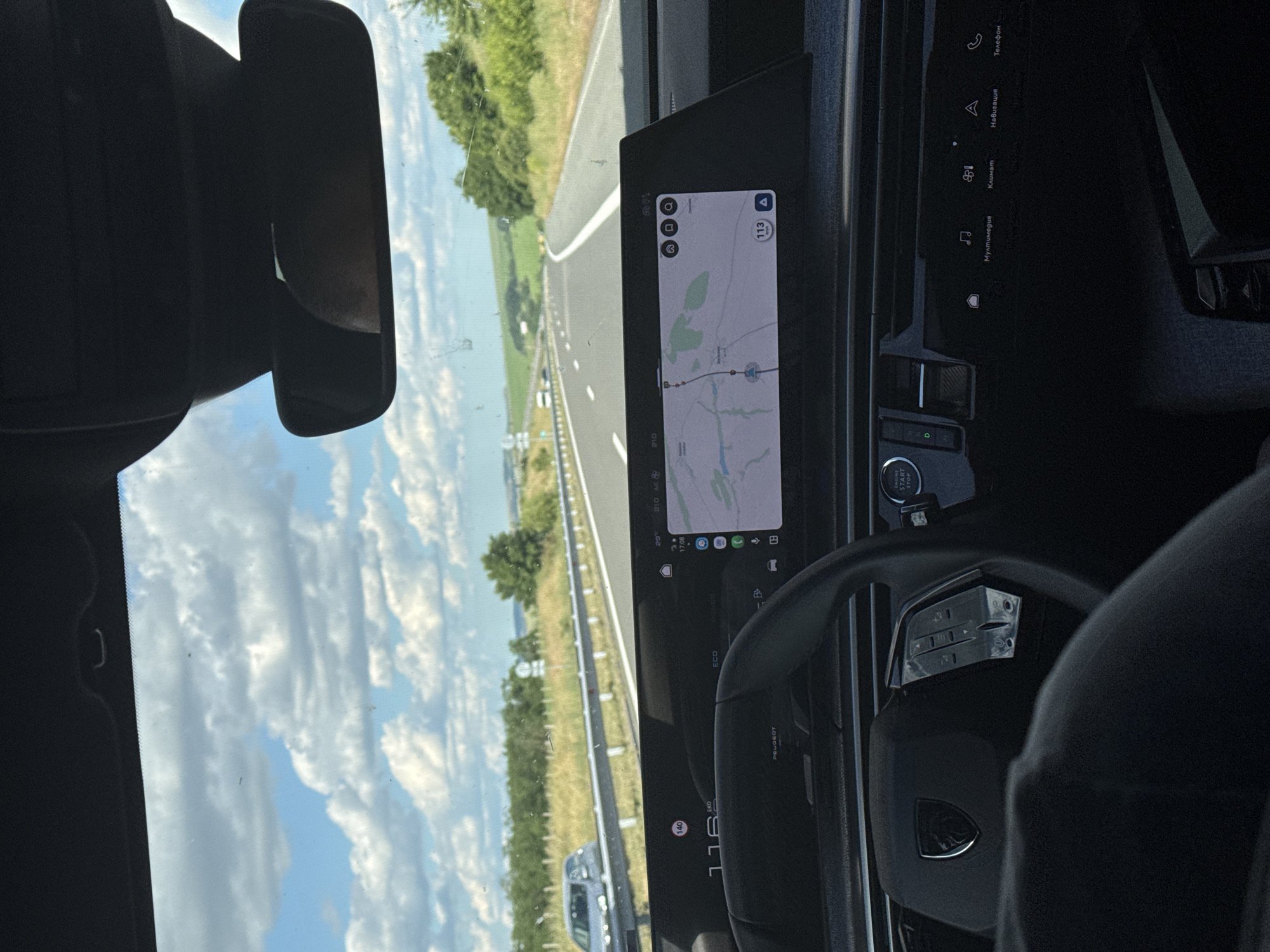 The image shows the interior of a car driving on a highway on a sunny day. The car is equipped with a large digital display screen showing navigation information. The sky is blue with scattered white clouds. The road ahead curves to the right. The surrounding landscape consists of green fields and trees. The car's steering wheel and dashboard are visible in the foreground. The speed limit sign shows 140. The current speed is 116 km/h. The navigation shows the car's current location with a red marker. The screen also displays icons for multimedia, climate control, navigation, and phone. The location is the highway near Zheleznik, Bulgaria.