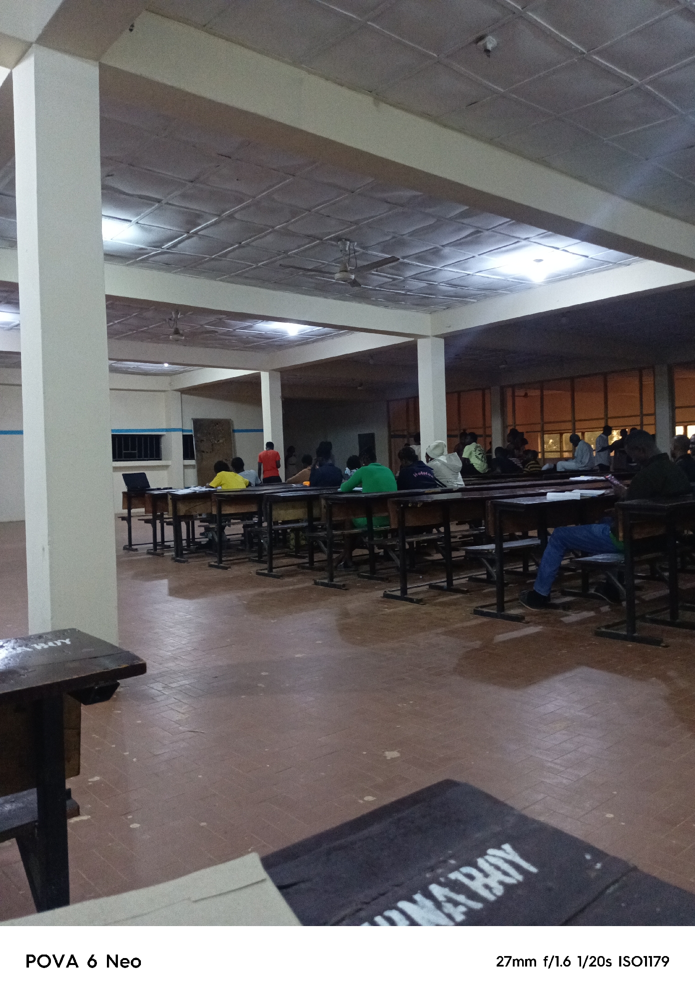 An indoor classroom scene is captured, likely at night or in low light, indicated by artificial lighting and the dark windows showing a dimly lit exterior. The room is furnished with rows of wooden desks and chairs, typical of a lecture hall or examination setting. Several people are seated at the desks, facing forward as if engaged in an activity such as studying or taking a test. Some are wearing casual clothing, while others have head coverings. A laptop is visible on one of the desks, and papers are scattered on some surfaces. The floor is tiled with a reddish-brown material, and the ceiling has a grid pattern with embedded lights and ceiling fans. The overall atmosphere is subdued, with the focus on the seated individuals and the functional classroom environment. The text "POVA 6 Neo" is present at the bottom left, and "27mm f/1.6 1/20s ISO1179" is at the bottom right, indicating camera settings and the device used.