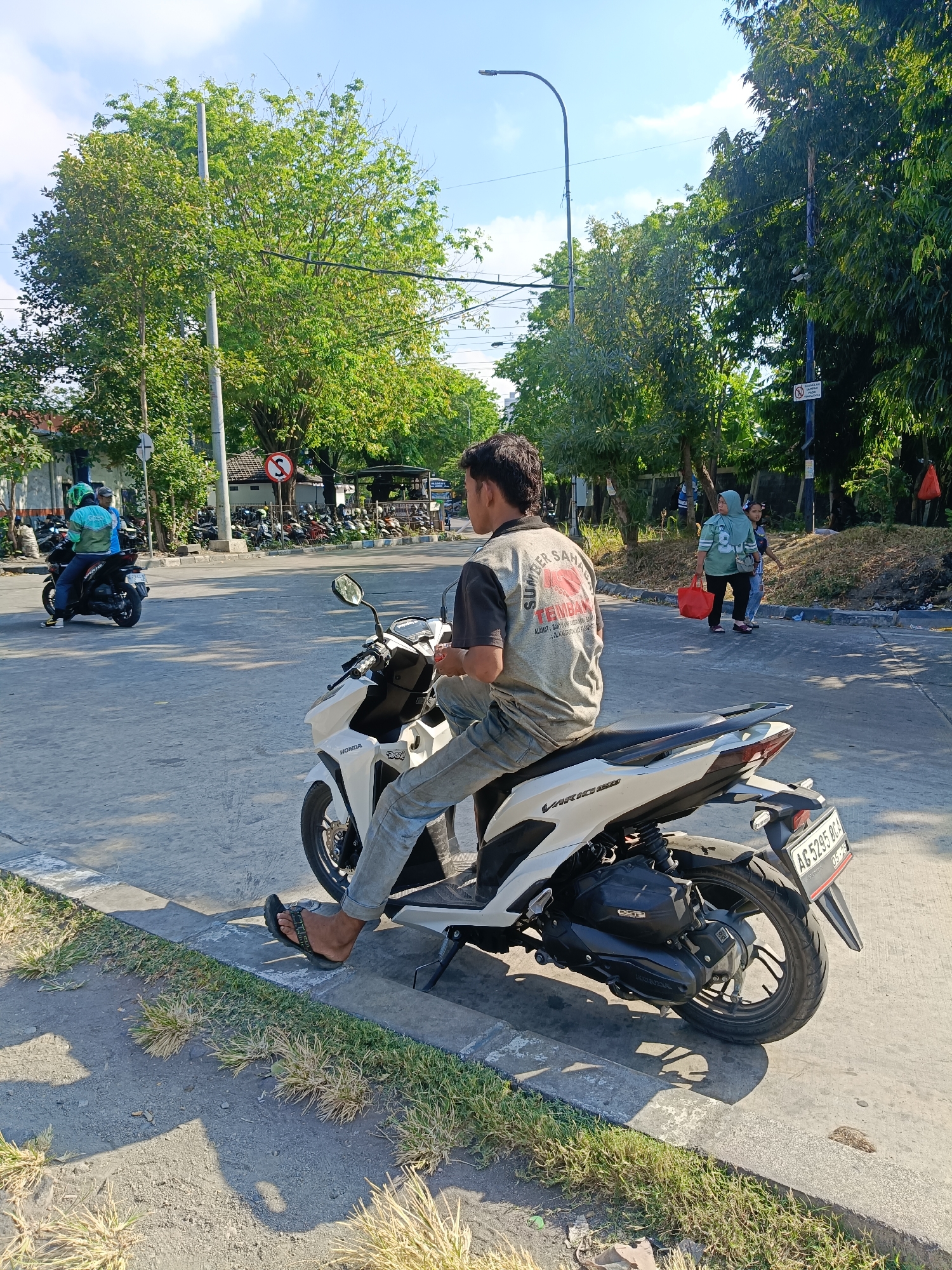 In the city of Kedungrejo, Indonesia, a man is seated on a white Honda Vario 150 motorcycle, facing away from the camera. He is wearing a grey t-shirt with red lettering that says "SUMBER SAH" and blue jeans. The motorcycle has a license plate reading "AG 5295 BCA". The man is wearing black flip-flops. The motorcycle is parked on a paved road with a concrete curb and some dry grass on the side. In the background, there are trees, a street light, a building, and several other motorcycles parked. Two women are walking in the background, one carrying a red bag. Another motorcycle with a rider wearing a green helmet is moving away from the camera on the left side of the frame. The sky is clear and blue.