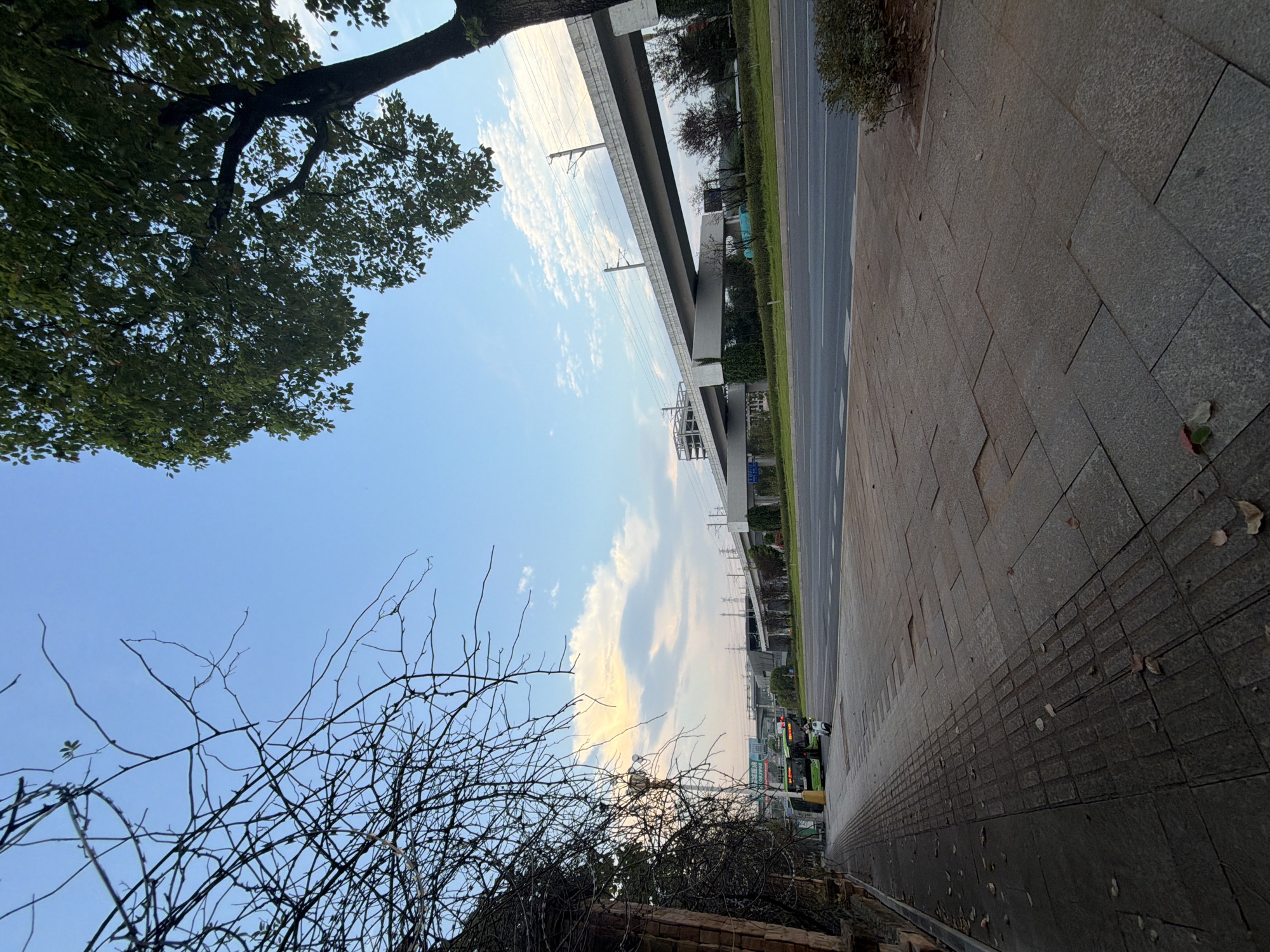 In Jiangjin, China, a paved sidewalk runs alongside a multi-lane road. On the left, bare tree branches and a brick wall are visible. Across the road, an elevated structure, likely for transportation, spans the scene. Behind this structure, a clear blue sky with scattered white clouds dominates the upper portion of the image. Several green buses and a white scooter are parked on the side of the road, near the sidewalk. The paving stones on the sidewalk are uneven, with some appearing to be missing or displaced. Trees with green leaves are present on the right side of the image, with one large tree's trunk and branches extending into the frame. The lighting suggests it is either early morning or late afternoon.