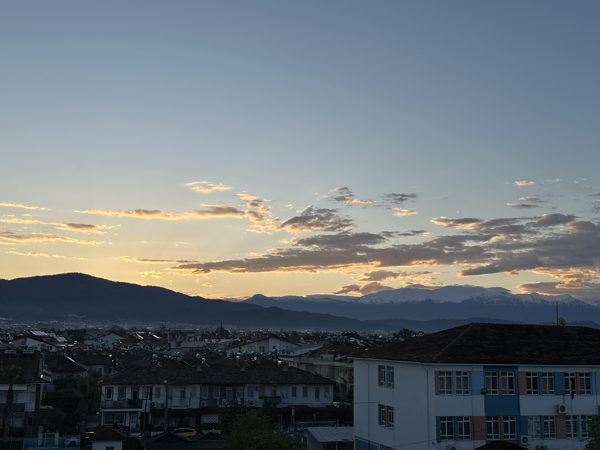 The image shows a cityscape in Fethiye, Turkey, at dusk. In the foreground are residential buildings with terracotta roofs, a light-colored school building with blue, pink, and white stripes on the right side, and trees. In the midground is a cluster of buildings and then a mountain range. The background features a sky with golden and gray clouds, suggesting a sunset. The mountains in the background have snow-capped peaks. The location is likely an elevated position within the city, providing a panoramic view.