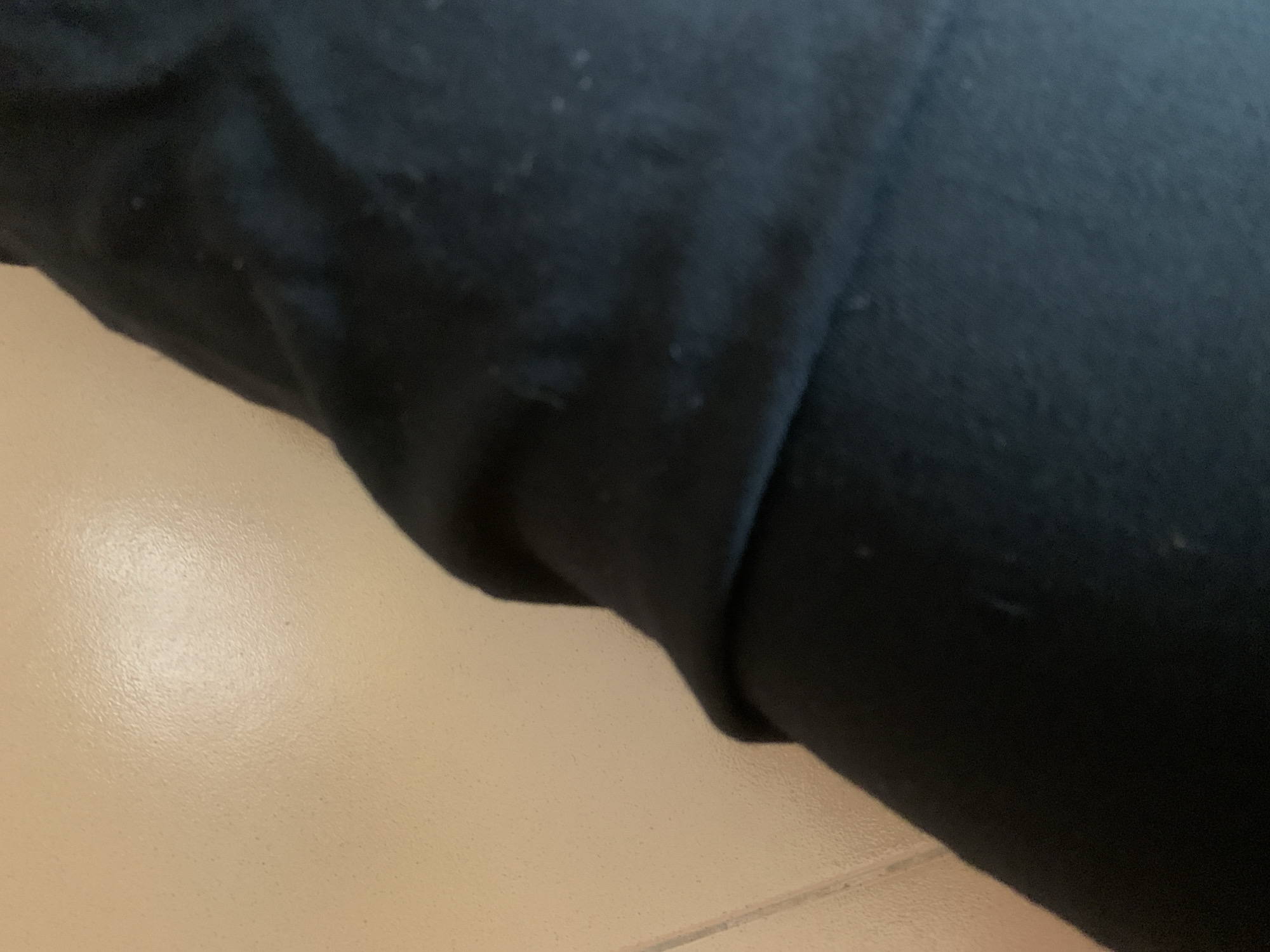 The image shows a close-up of a person's black clothing, likely pants or leggings, against a pale, textured wall. The fabric of the clothing is draped and folded, creating shadows and highlights that reveal its texture. There are a few small specks or imperfections visible on the black fabric. The wall appears to be painted a light beige or cream color and has a slightly uneven, stucco-like finish. The lighting suggests an indoor environment, with a soft, diffused light source coming from the upper left. There is no indication of the city of Jauro Yinu, Nigeria, or any specific geographical features.
