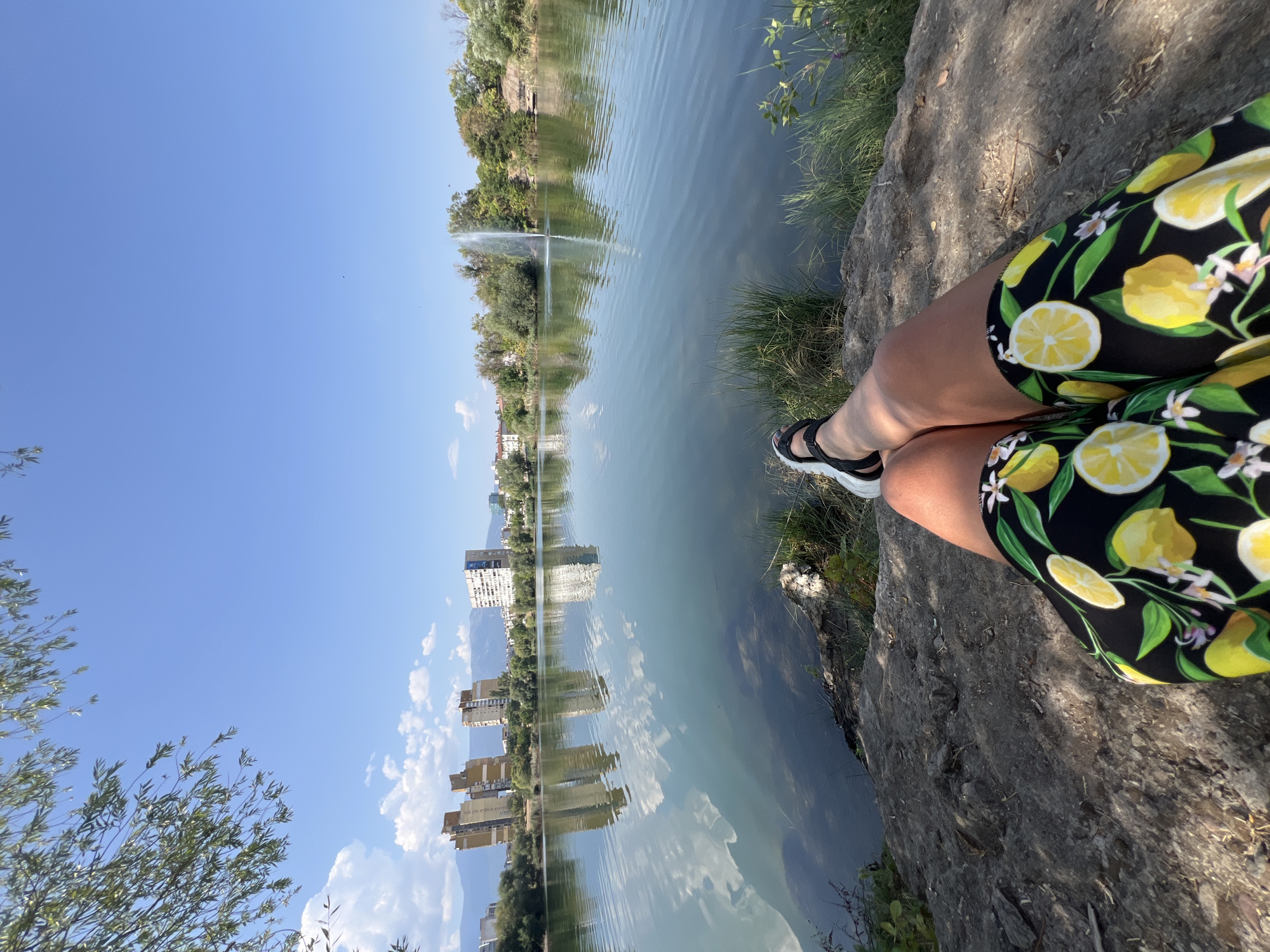 The image shows a first-person perspective of someone sitting on a rocky bank overlooking a lake. The person is wearing black shorts with a pattern of yellow lemons and white flowers, and black sandals. The lake has calm water with reflections of the sky and buildings on the opposite shore. In the distance, there are several modern apartment buildings and mountains. A fountain is visible in the middle of the lake, spraying water upwards. Lush green trees and vegetation surround the lake. The sky is a clear blue with scattered white clouds. This scene is likely from one of Sofia's parks that feature lakes, such as the Lake Ariana park in Druzhba district, or the park around the National Palace of Culture.