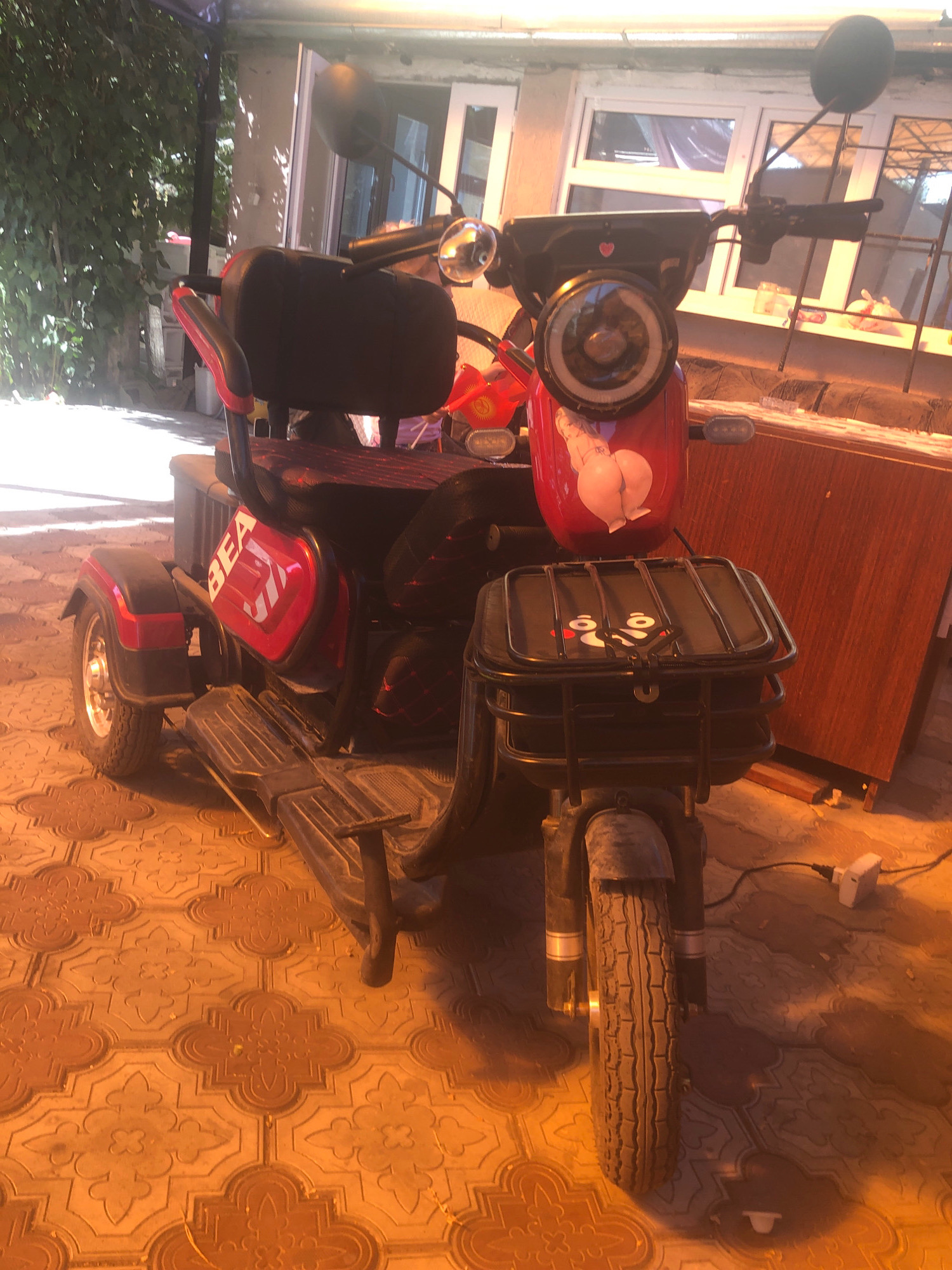 An electric three-wheeled scooter is parked on a patterned tile surface, possibly in a courtyard or driveway. The scooter is red and black, with a black seat and a basket on the front. There is a sticker of a woman's buttocks on the front of the scooter. A wooden cabinet or shelf is visible behind the scooter. In the background, there is a building with windows and greenery. The scene is illuminated by sunlight, casting shadows. The location is Bishkek City, Kyrgyzstan.