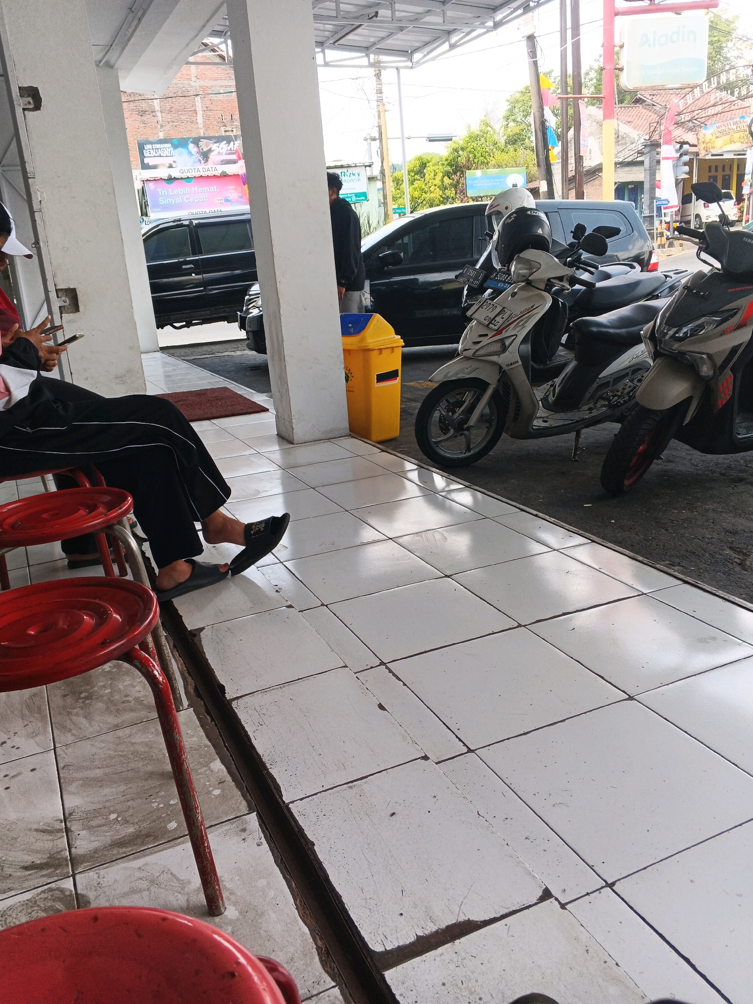 The image captures a street scene in Genuk Barat, Indonesia, specifically outside a building with white pillars and a tiled floor. In the foreground, two red metal stools are visible, one in focus and another partially out of frame. A person is seated on one of the stools, wearing black trousers and black sandals, holding a mobile phone. To the right of the person, a yellow trash can stands next to a row of parked motorbikes. The motorbikes, including a white Yamaha and another partly visible, are lined up in front of a black SUV. In the background, various shops and buildings are visible, along with trees and a street sign for "Aladin". The overall environment suggests a commercial area, possibly a waiting area or a roadside establishment.