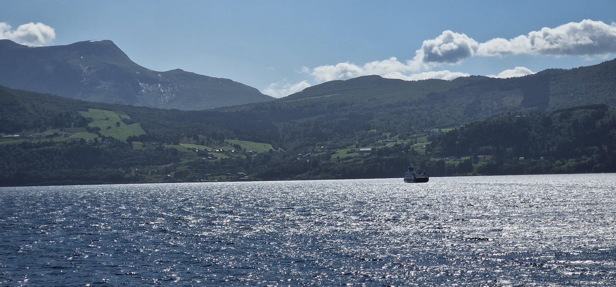 This image depicts a scenic view of the Volda Fjord in Volda, Norway, on a sunny day. The foreground is dominated by the dark blue water of the fjord, sparkling with sunlight, creating a shimmering effect. In the middle ground, a ferry is visible, moving across the water. The background features a lush, green mountainside dotted with small houses and farms, characteristic of the rural landscape of Volda. The upper portion of the image showcases a clear blue sky with a few fluffy white clouds. The overall impression is one of tranquility and natural beauty.