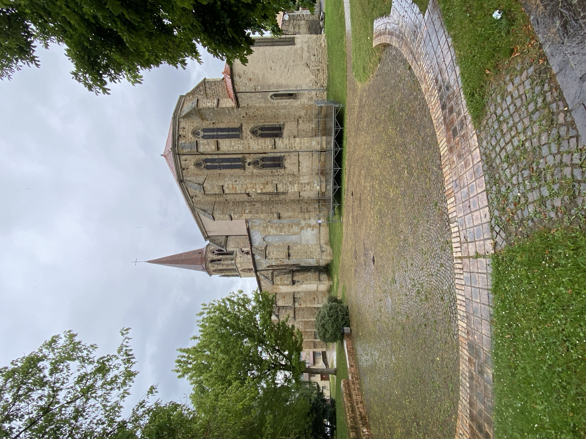 The image displays a historic stone church or cathedral, likely in Billom, France, viewed from an outdoor park or courtyard. The large ecclesiastical building features robust stone walls, a reddish-brown tiled roof, and several arched windows, some with decorative tracery. A tall, slender spire, topped with a cross, rises from one end of the structure, adding to its architectural grandeur. Metal scaffolding is visible along a section of the church's side wall, suggesting ongoing maintenance or restoration.
The ground surrounding the church consists of a wide, damp cobblestone path, which appears to have a curved edge made of larger, rectangular pavers. Lush green grass, dotted with small white flowers in some areas, abuts the paved sections. Several deciduous trees with dense green foliage are prominent in the foreground and along the periphery of the scene, providing a natural frame. The sky is uniformly overcast and grey, indicating cloudy weather and a calm atmosphere. The damp appearance of the ground and foliage suggests recent rainfall. No people or visible text are present in the image.