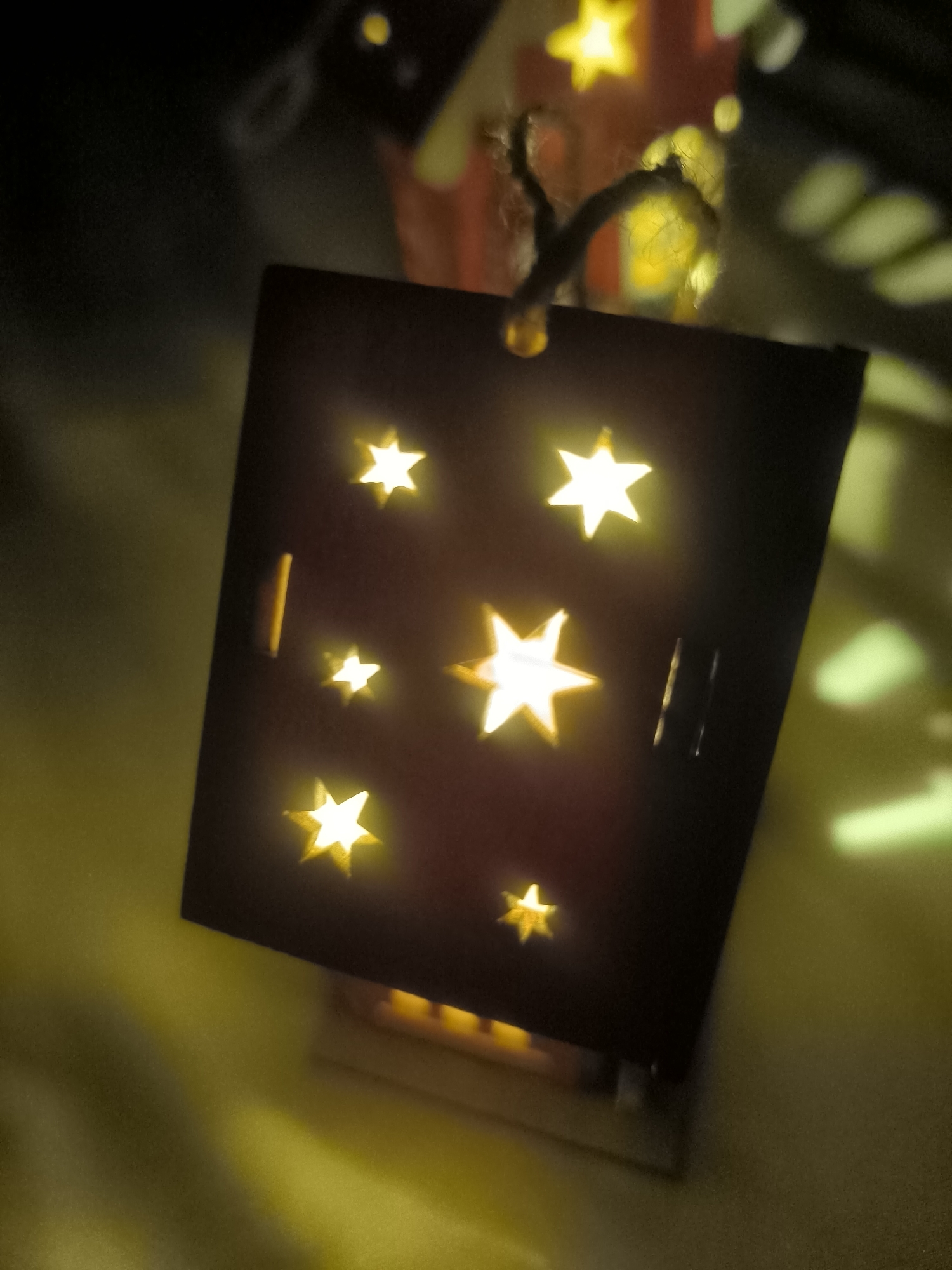 The image shows a close-up, low-angle shot of a decorative lantern with cut-out star shapes, illuminated from within by a warm yellow light. The lantern is dark brown or black and hangs by a rough twine. Several stars are visible on the front surface of the lantern, some larger and brighter than others. To the left of the lantern, the background is a blurry, soft yellow. To the right, there are out-of-focus shapes and light patterns that suggest a background environment with other illuminated objects or reflections. The overall impression is one of a cozy, possibly festive, atmosphere. The location is not discernible from the image.