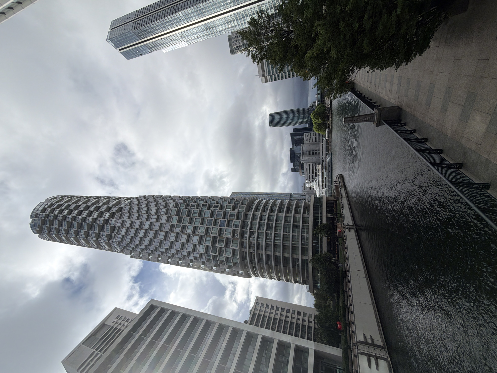 The image presents an urban waterside landscape, captured from a low, upward-looking perspective and rotated approximately 90 degrees clockwise. A prominent, tall, cylindrical skyscraper with a distinctive, intricately patterned facade of numerous square windows and curved balconies dominates the mid-ground, extending from the lower left towards the upper middle of the frame. Above it, another sleek, modern skyscraper with a glass and metal exterior is partially visible, extending out of the top of the frame.
A dark, rippled body of water, resembling a canal or dock, flows horizontally across the right side of the image. It is bordered by a paved pedestrian walkway of light grey flagstones, featuring a dark metal railing or fence. Midway along this waterway, a small, block-like structure with a vertical element extends into the water from the walkway. Several green trees are visible along the banks and interspersed among the buildings, adding natural elements to the urban setting.
The sky is heavily overcast with varying shades of grey and white clouds, indicating a cloudy daytime. Other contemporary buildings, predominantly light-colored with angular and slatted designs, are visible in the background and foreground, contributing to the modern metropolitan atmosphere. No people or discernible activities are visible, and there is no visible text. The architecture and waterside setting are characteristic of areas like Canary Wharf in London, United Kingdom.