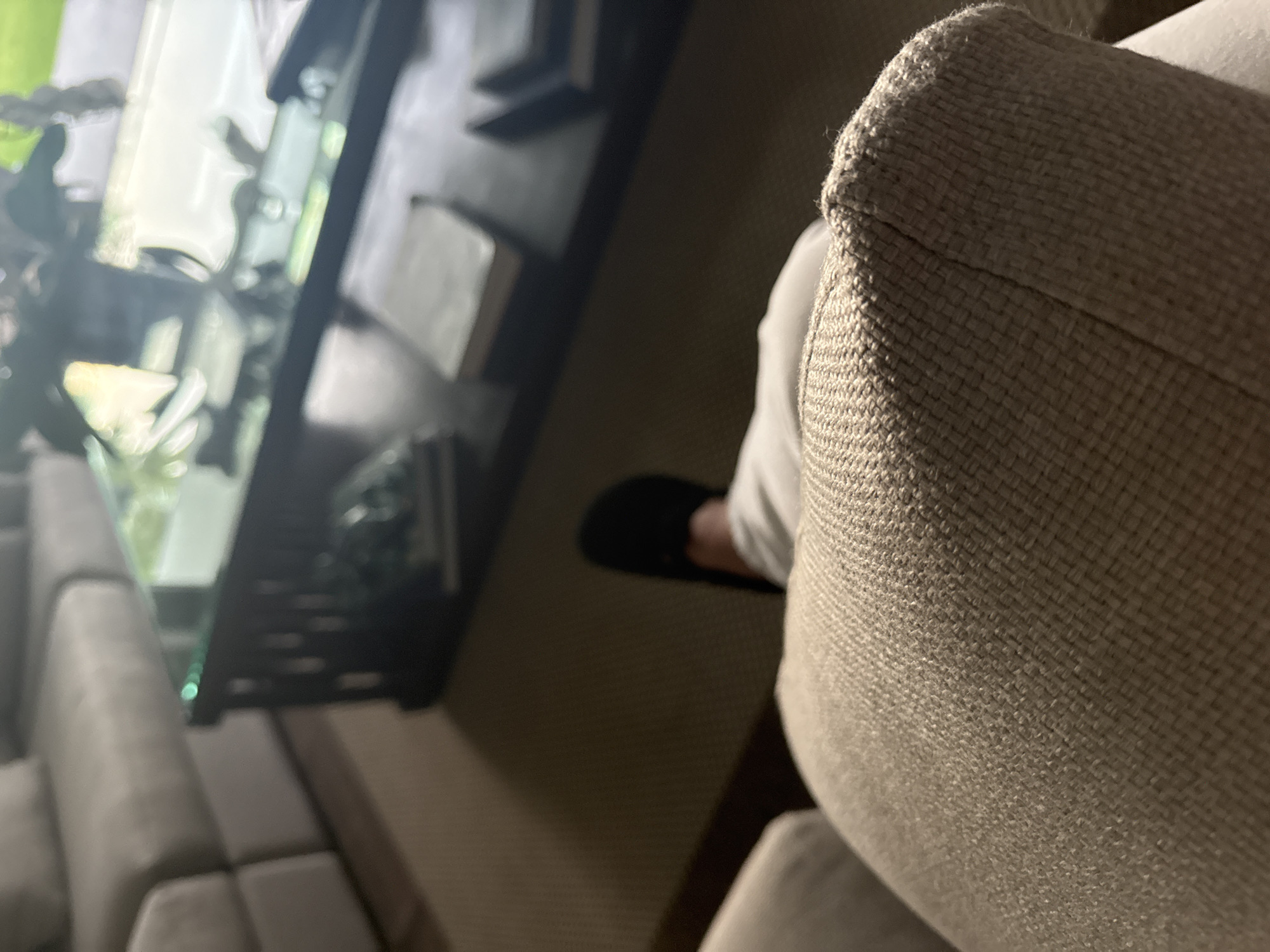 The scene is set indoors in a residential living area within San Bernardino Tlaxcalancingo, Mexico. The image presents a low-angle, first-person perspective. In the immediate foreground, a textured, woven, light-beige upholstered surface, likely a sofa armrest or cushion, occupies the lower portion of the frame. Directly beyond this, a human leg, clad in light-colored trousers, extends forward, with a black slipper or closed-toe shoe visible on the right foot. The individual appears to be seated. In the midground, a dark-colored coffee table with a reflective, possibly glass, top is situated. Objects, primarily appearing as books, are visible stacked on a lower shelf of the table. The reflective tabletop displays indistinct reflections of light sources and green foliage. In the background, a large, light-colored, multi-sectional sofa is partially visible on the left, and the floor is covered by a light-brown carpet. The lighting appears dim and ambient.